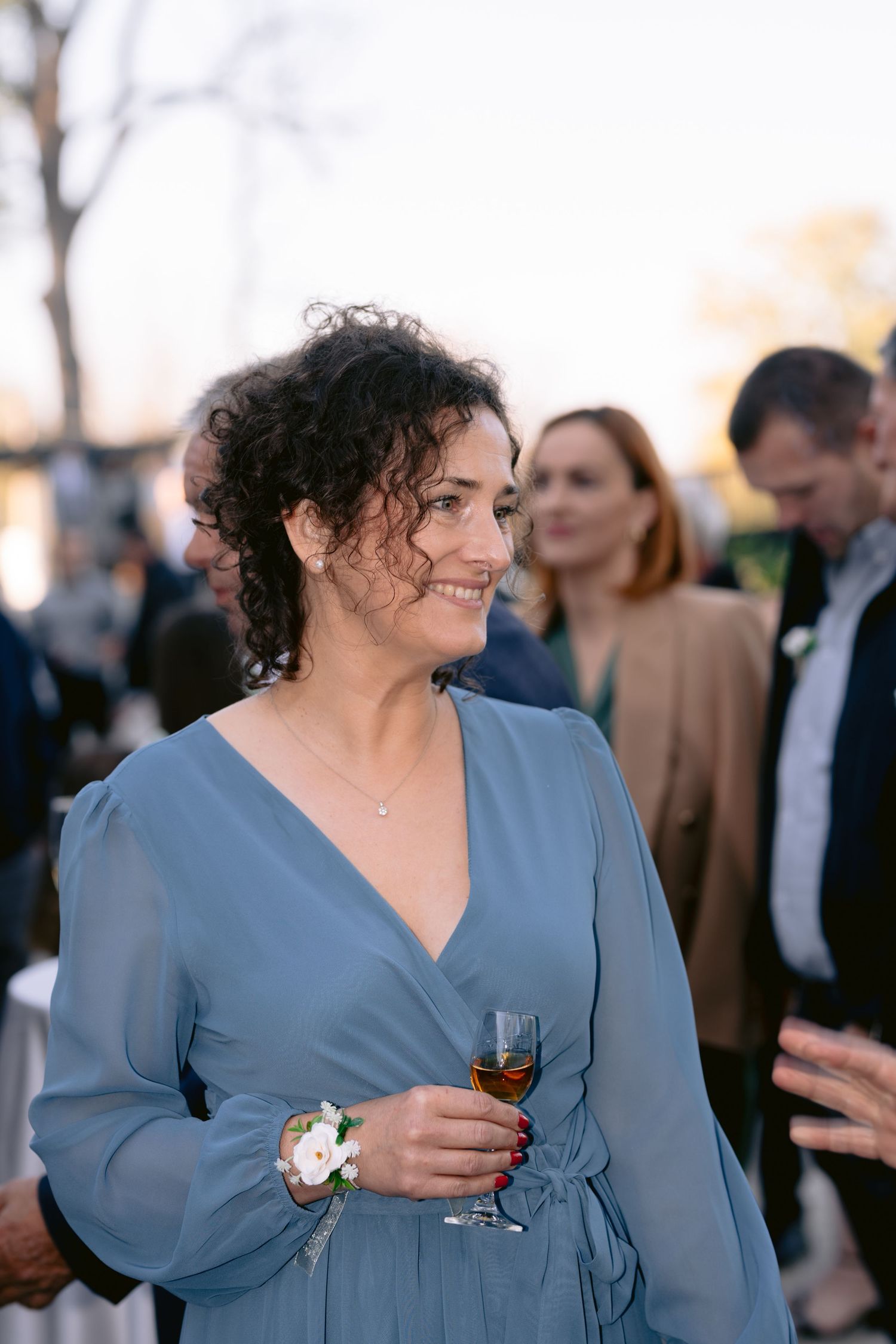 Woman in blue dress holding wine glass at outdoor gathering with guests in background.