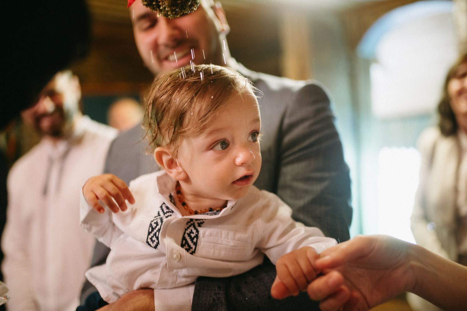 A baby in a white embroidered outfit being held by an adult in a gray suit at a festive indoor event.