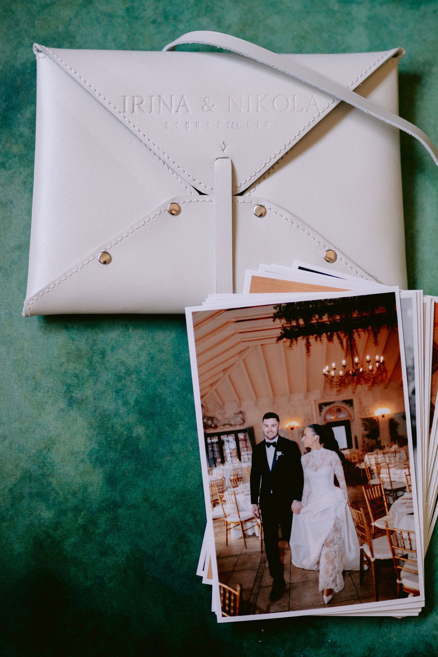 Bijela kožna pismo-torbica sa natpisom Irina i Nikola pored štampanih fotografija mladenaca koji šetaju u elegantnom prostoru Ville Slatine.