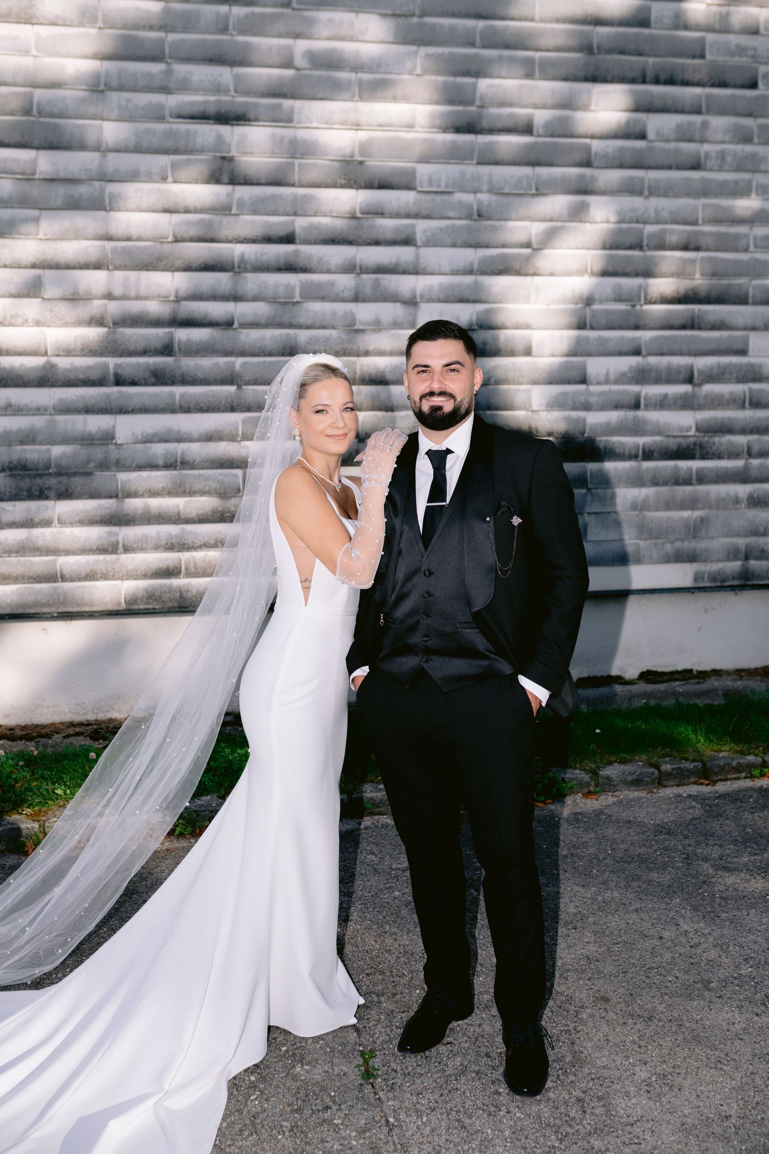 Bride in white gown with veil and groom in black tuxedo posing together outdoors against a gray brick wall.