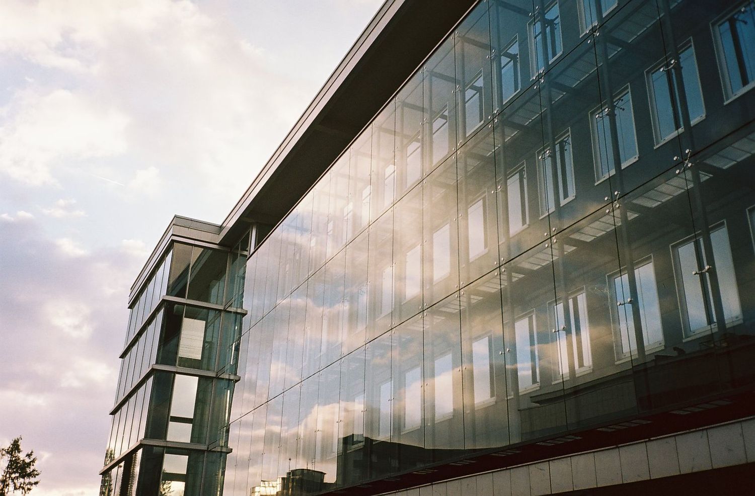 Modern glass office building facade reflecting clouds and sky at sunset with angular architectural details.