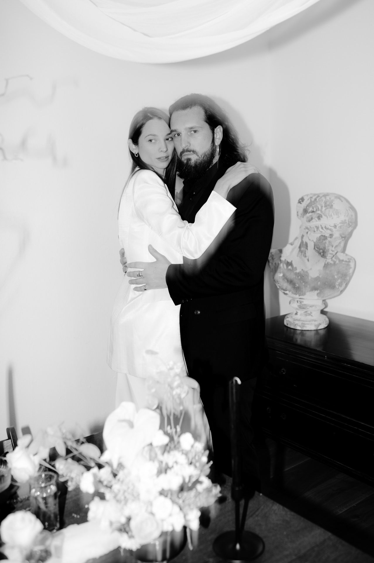 Black and white wedding photo of bride and groom embracing, with floral arrangements and a decorative bust in the background.