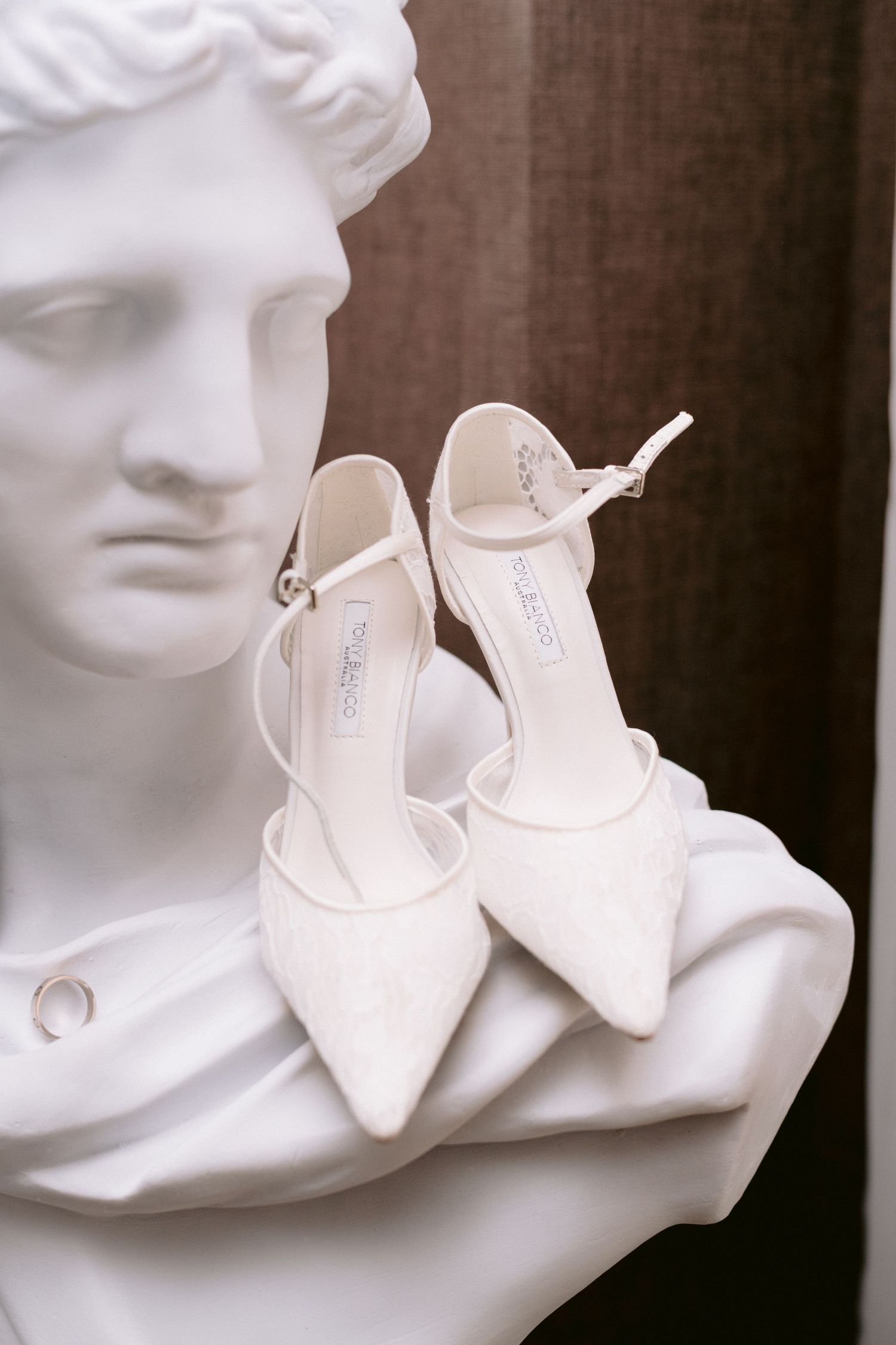 White bridal heels displayed on a classical marble bust sculpture, elegant wedding shoes with ankle strap detail.