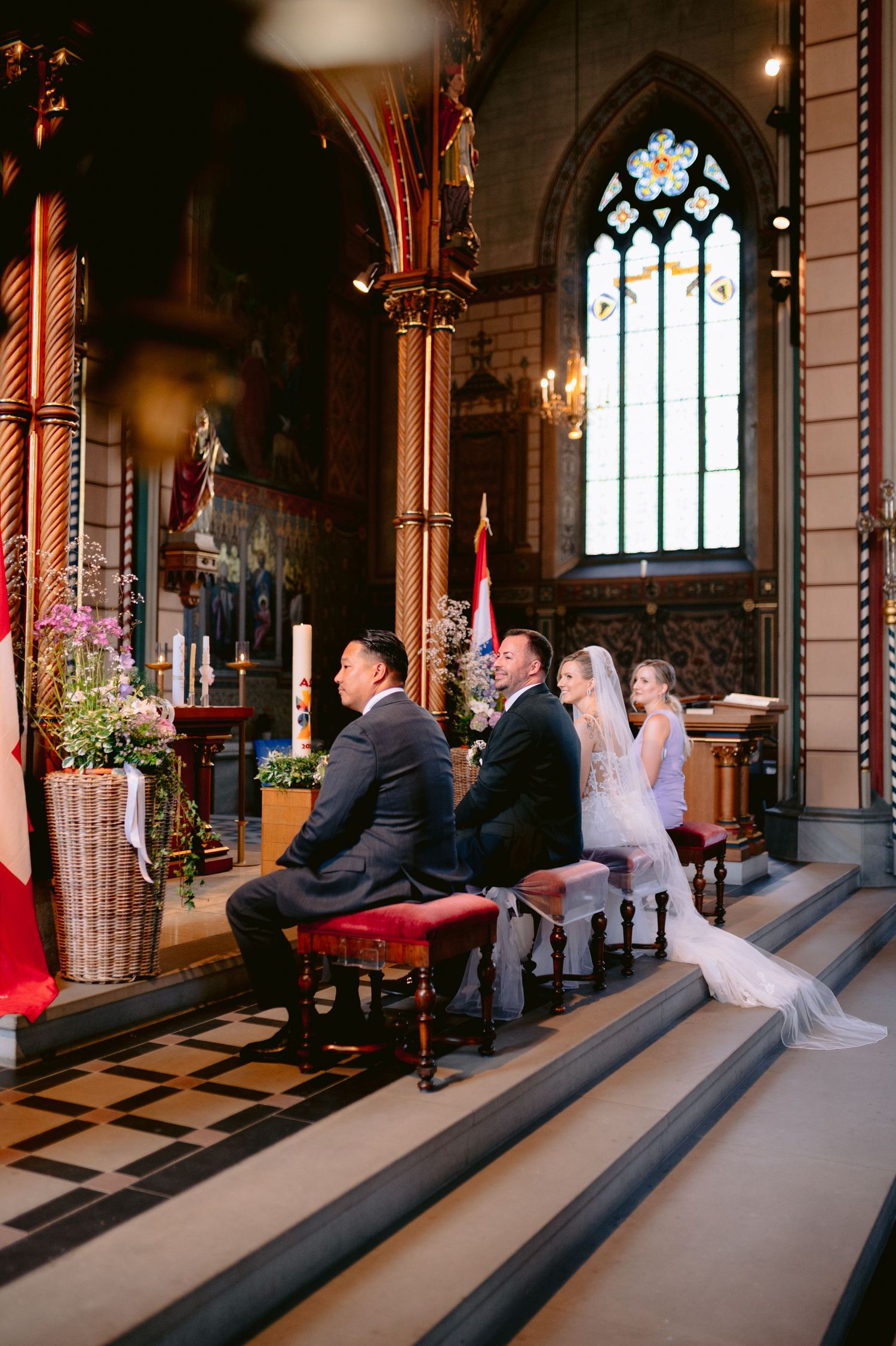 Bride and groom kneel at church altar during wedding ceremony in ornate Gothic chapel with stained glass windows.