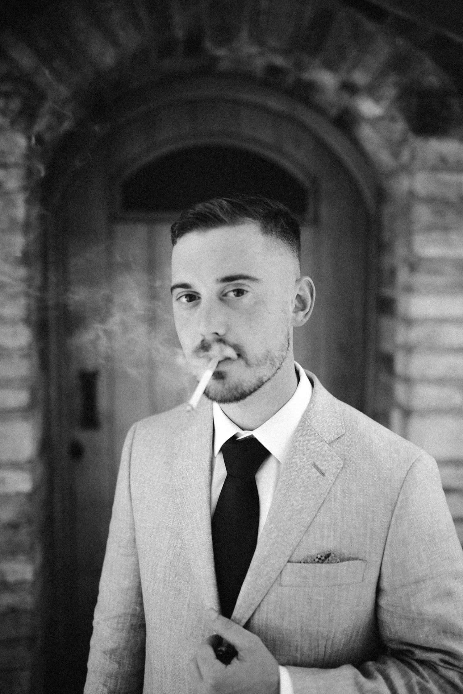 Black and white portrait of person in light suit smoking against stone archway.