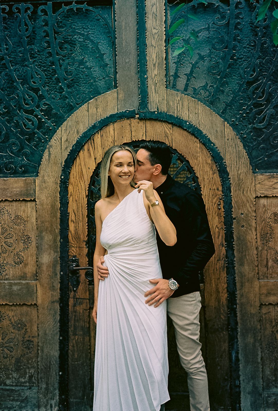 A couple embraces in front of a rustic wooden door with arched details and turquoise walls.