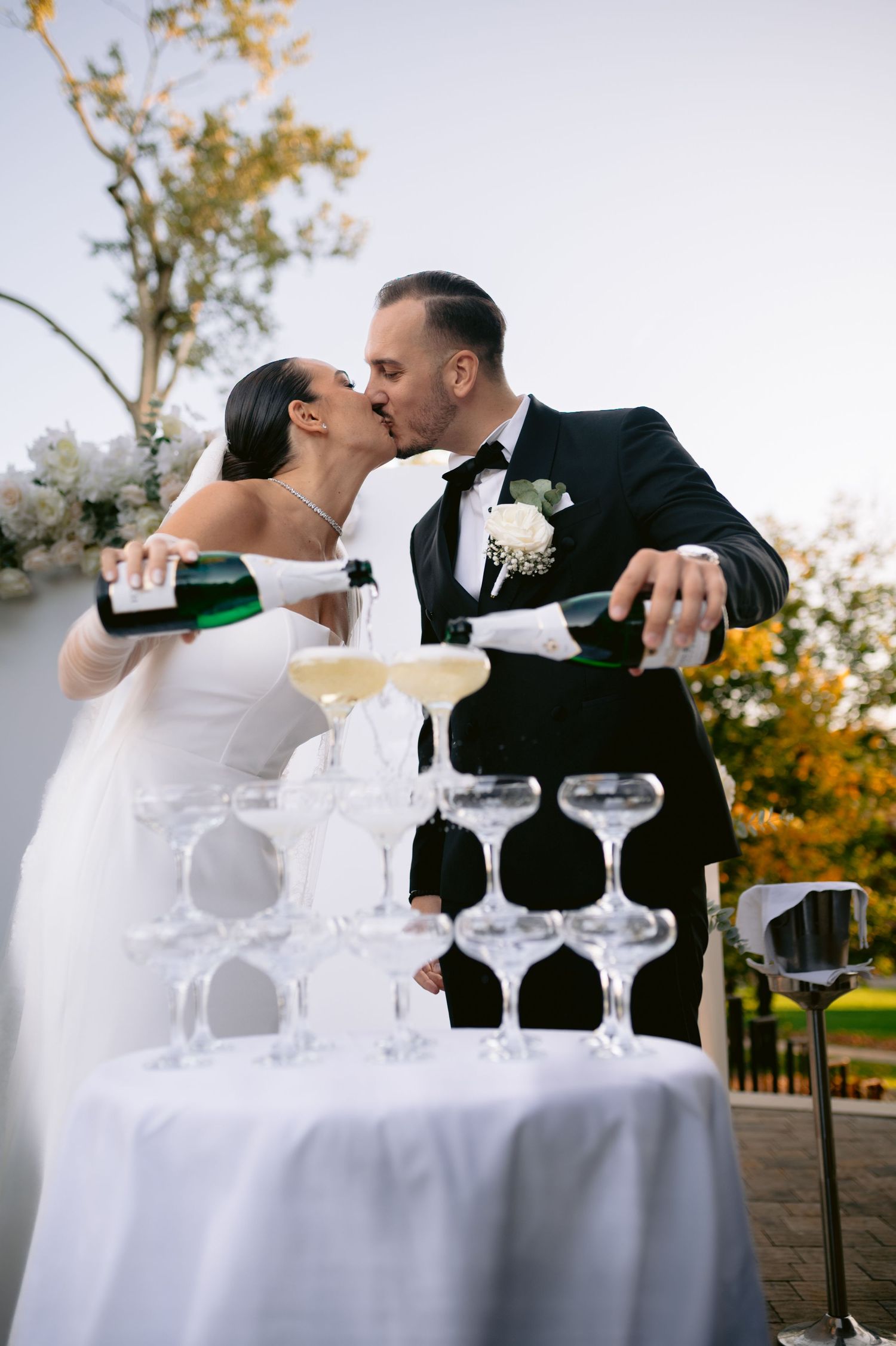 A romantic wedding couple shares a kiss behind champagne glasses arranged on a white tablecloth outdoors.