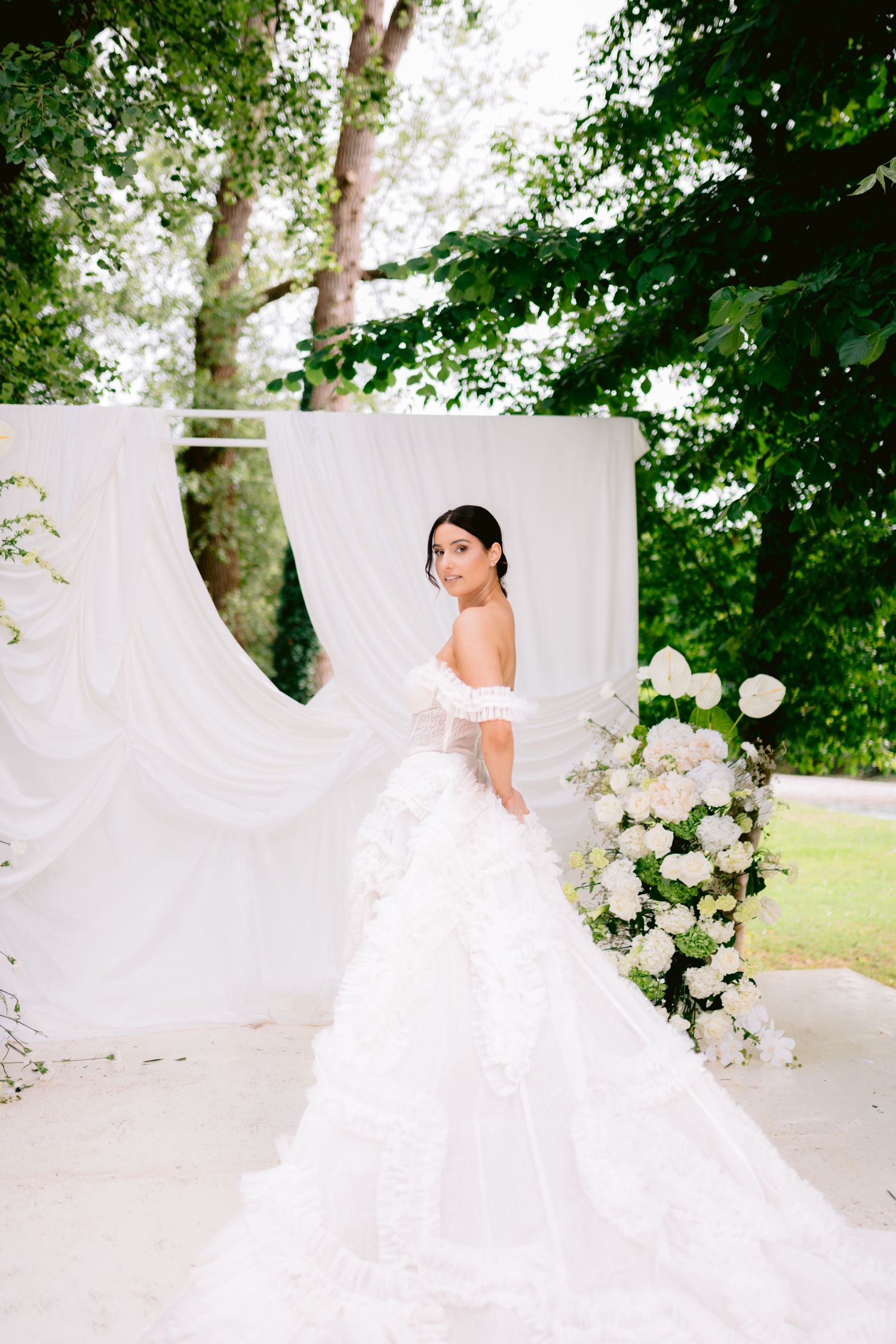 Elegant bride in off-shoulder ruffled gown poses before white draped arch with floral decor.