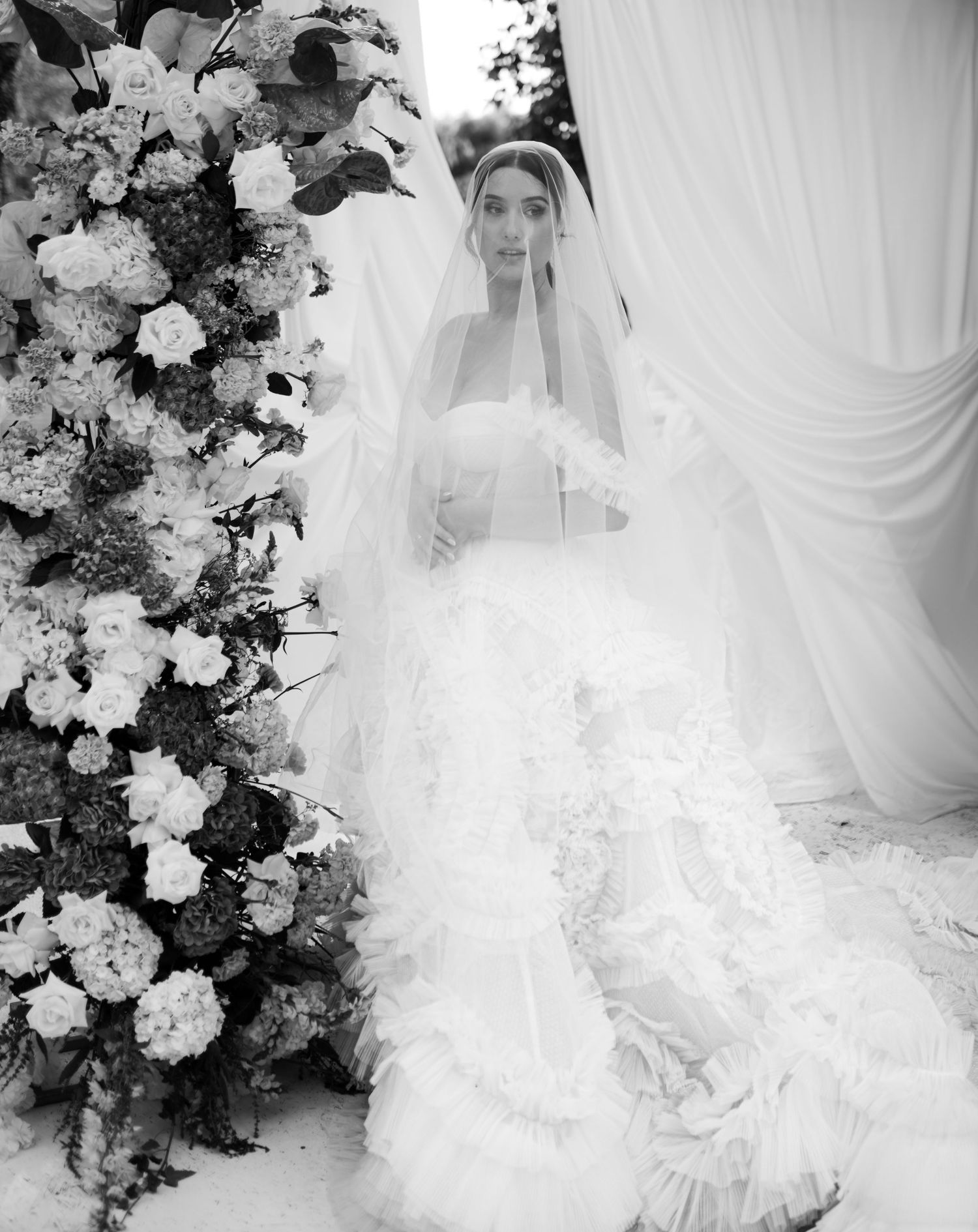Bride in a ruffled white wedding gown and veil poses beside a lush floral arrangement in a black and white photo.