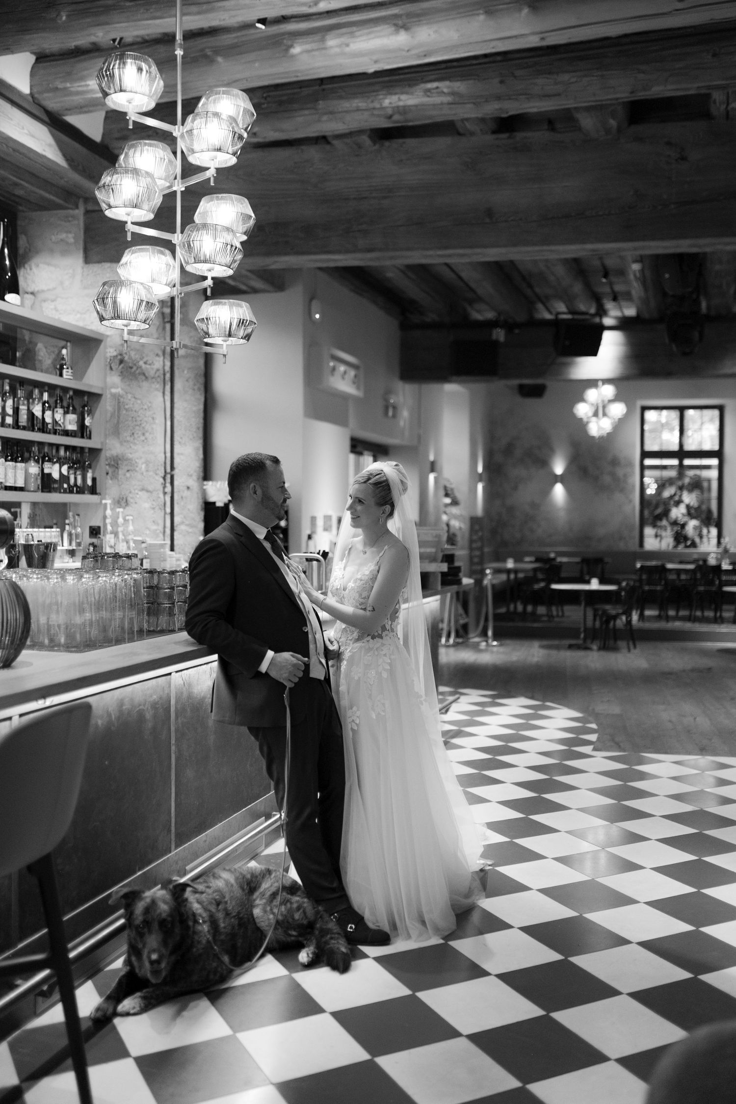 Bride and groom stand at bar with dog on checkered floor in black and white wedding photo.