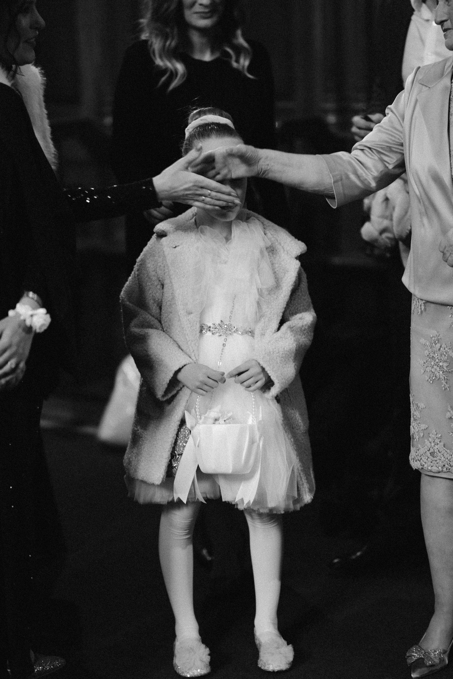 A black and white photo of a child in a white dress and veil being blessed during a religious ceremony.
