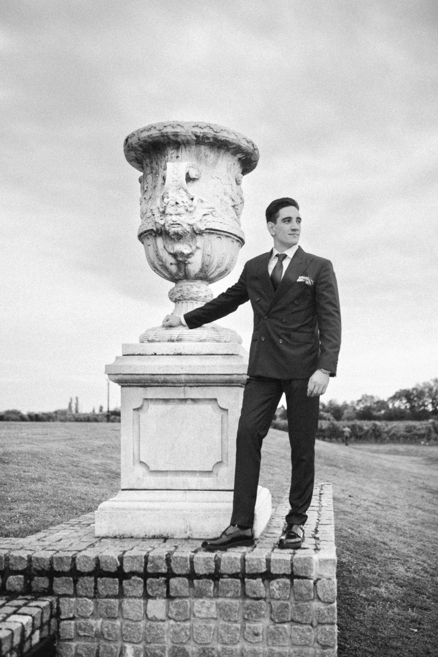 Black and white photo of a person in a suit standing next to an ornate classical stone urn on a brick pedestal.