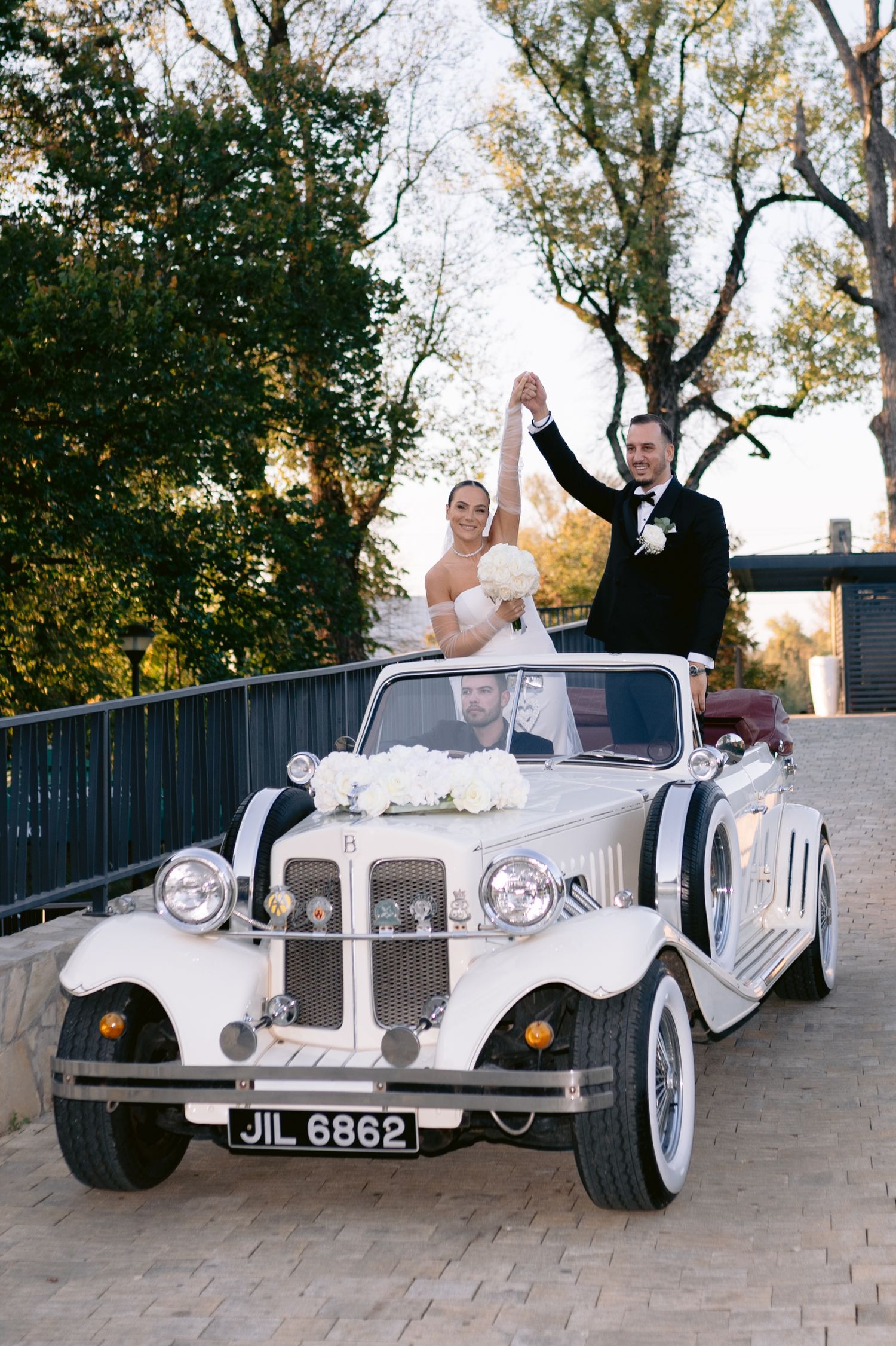 A happy couple waves from a vintage white wedding car on a bridge surrounded by bare winter trees in black and white.