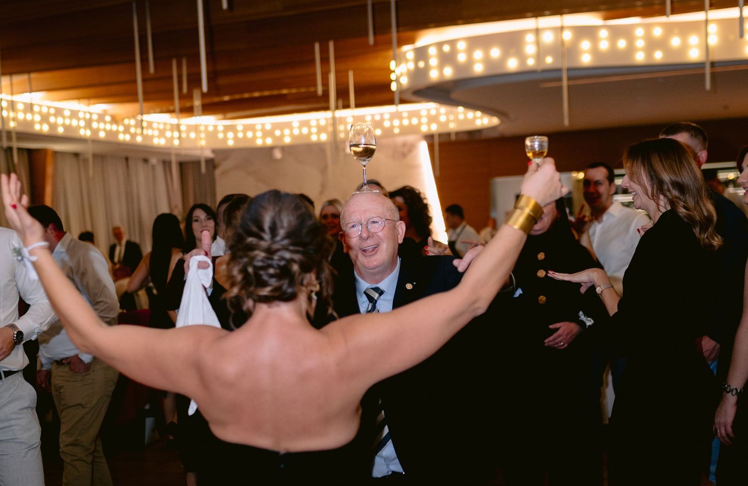 Guests celebrate and dance joyfully under circular chandeliers at a lively wedding reception.