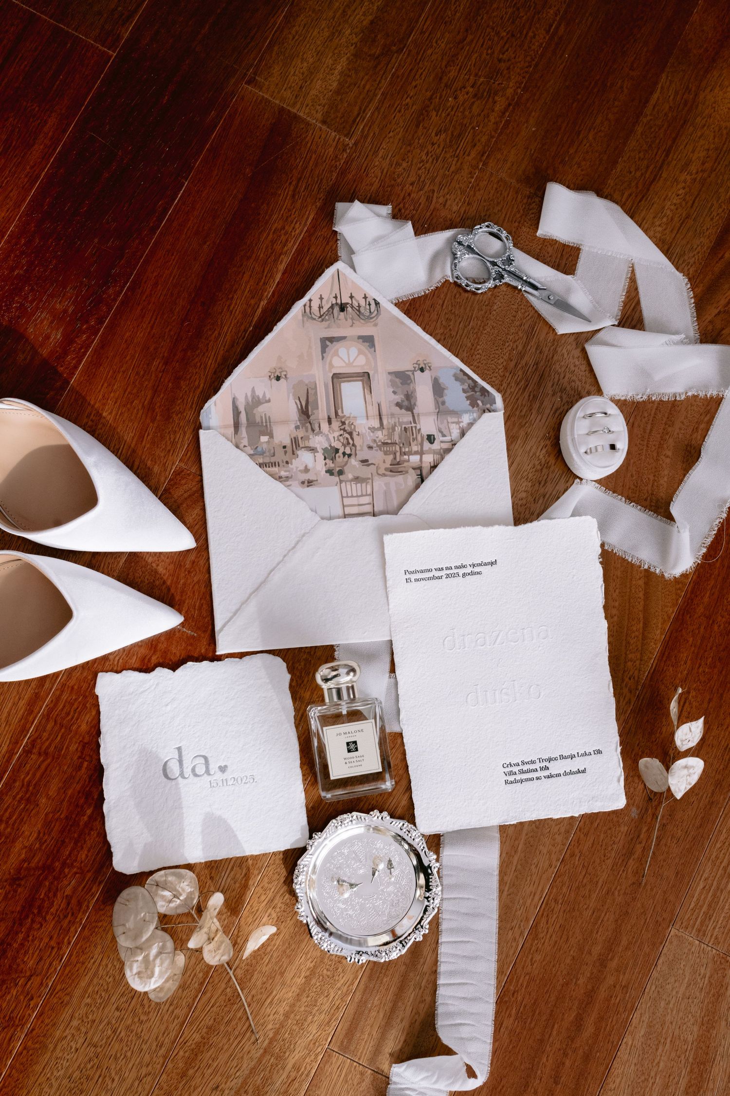 Elegant wedding flat lay with white heels, Jo Malone perfume, invitation suite, ribbon, and jewelry on wood floor.