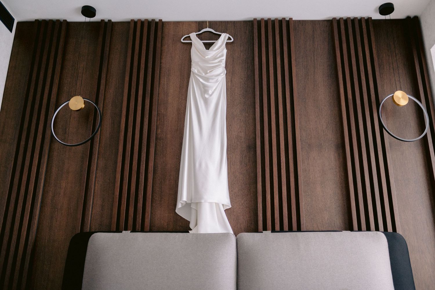 White wedding dress hanging on a wooden slatted wall between two gold ring sconces above a gray upholstered headboard.