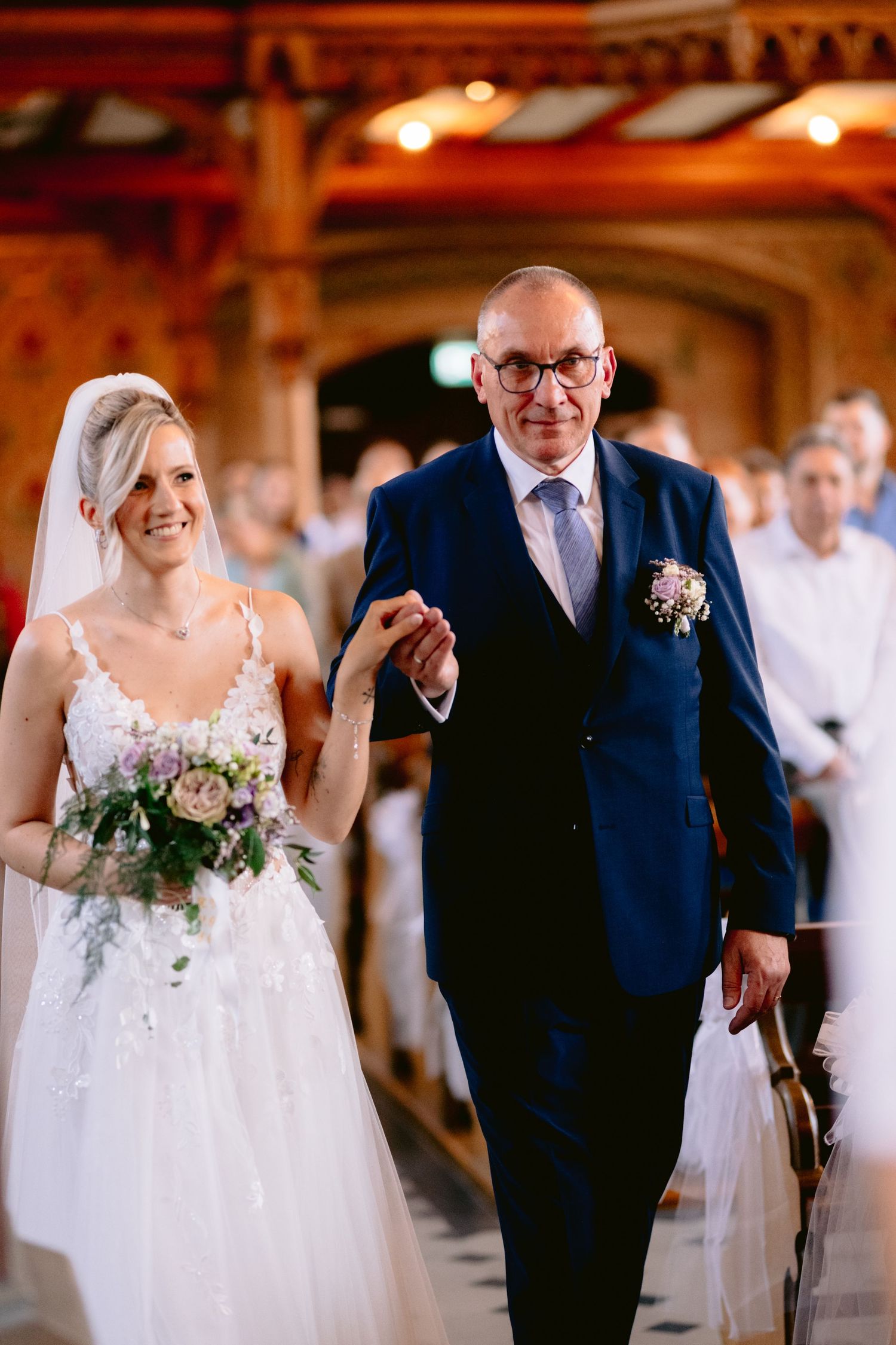 Bride holding bouquet walks down church aisle with older man in navy suit during wedding ceremony.