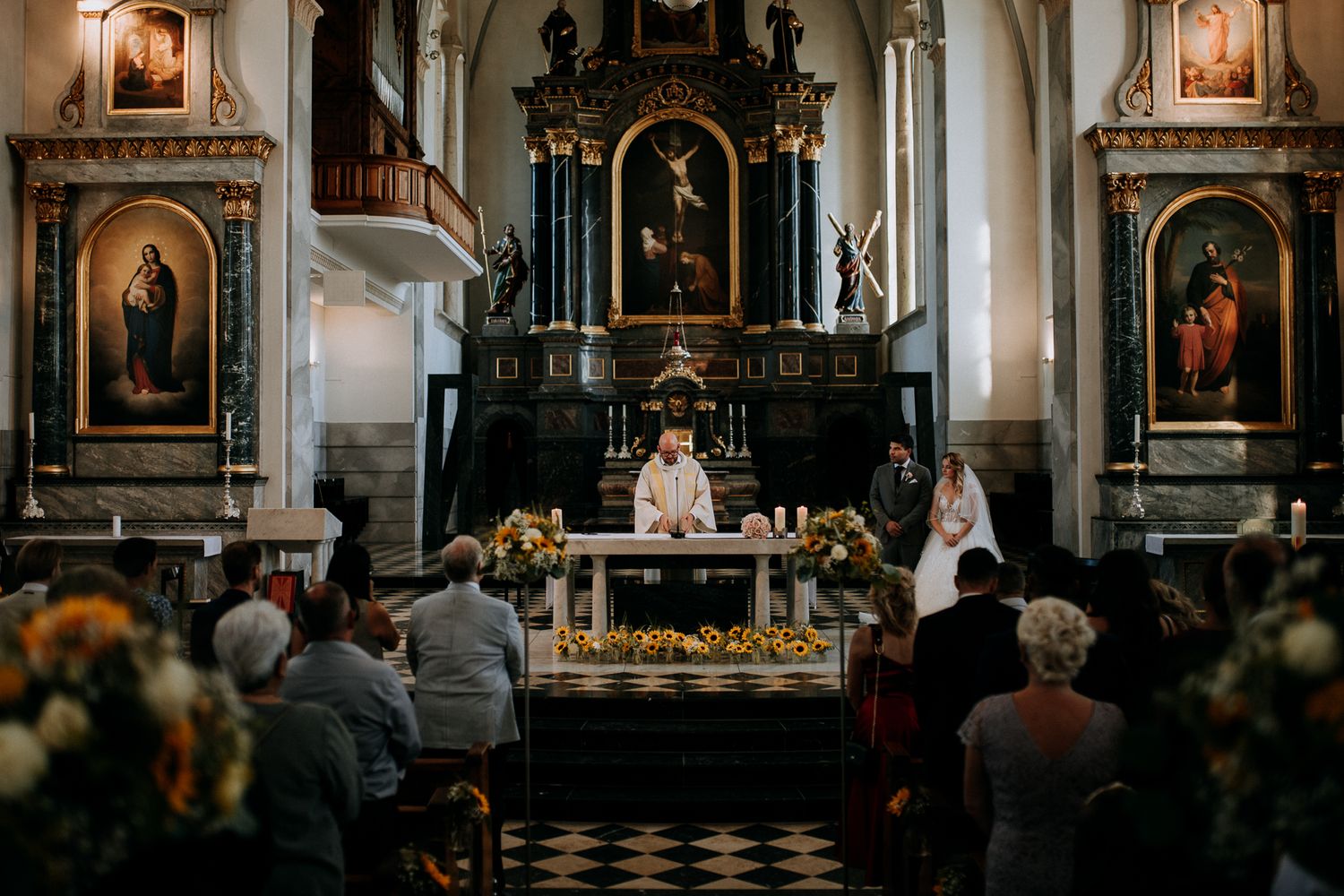 Elegant church wedding ceremony with couple at ornate altar, surrounded by floral arrangements and guests in historic baroque church.