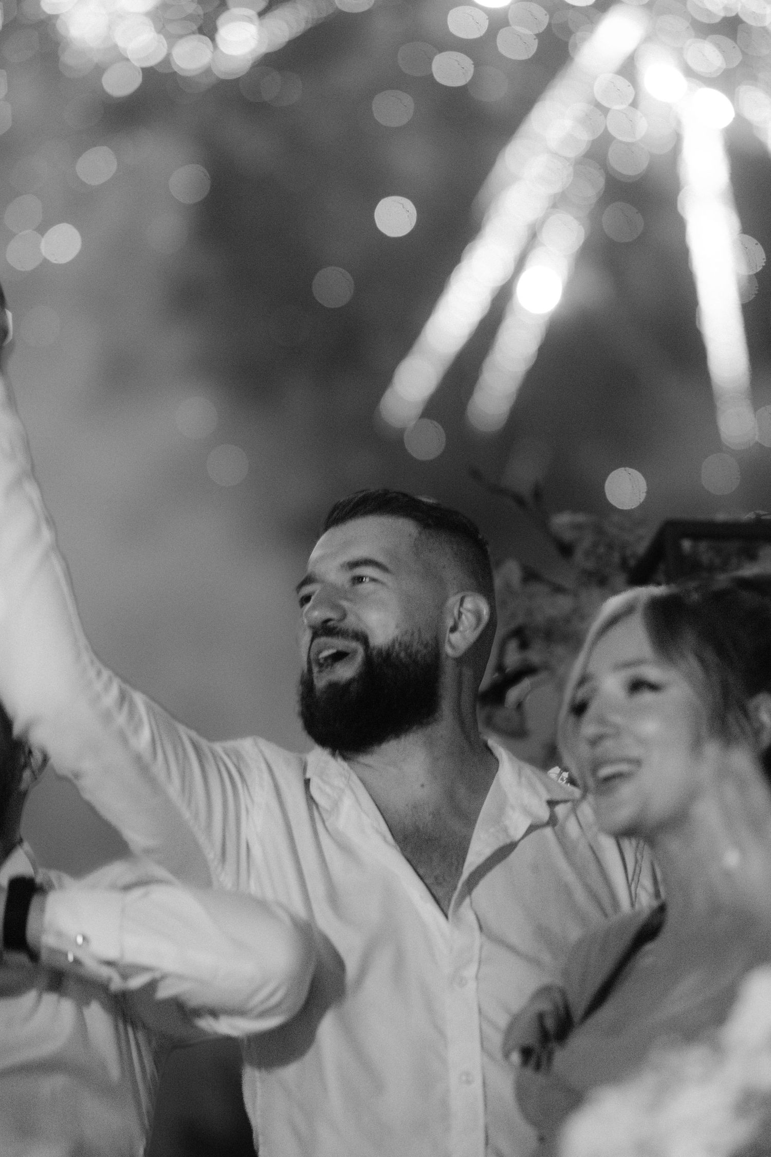 Black and white photo of a bearded man in white shirt smiling at fireworks display with a woman beside him.