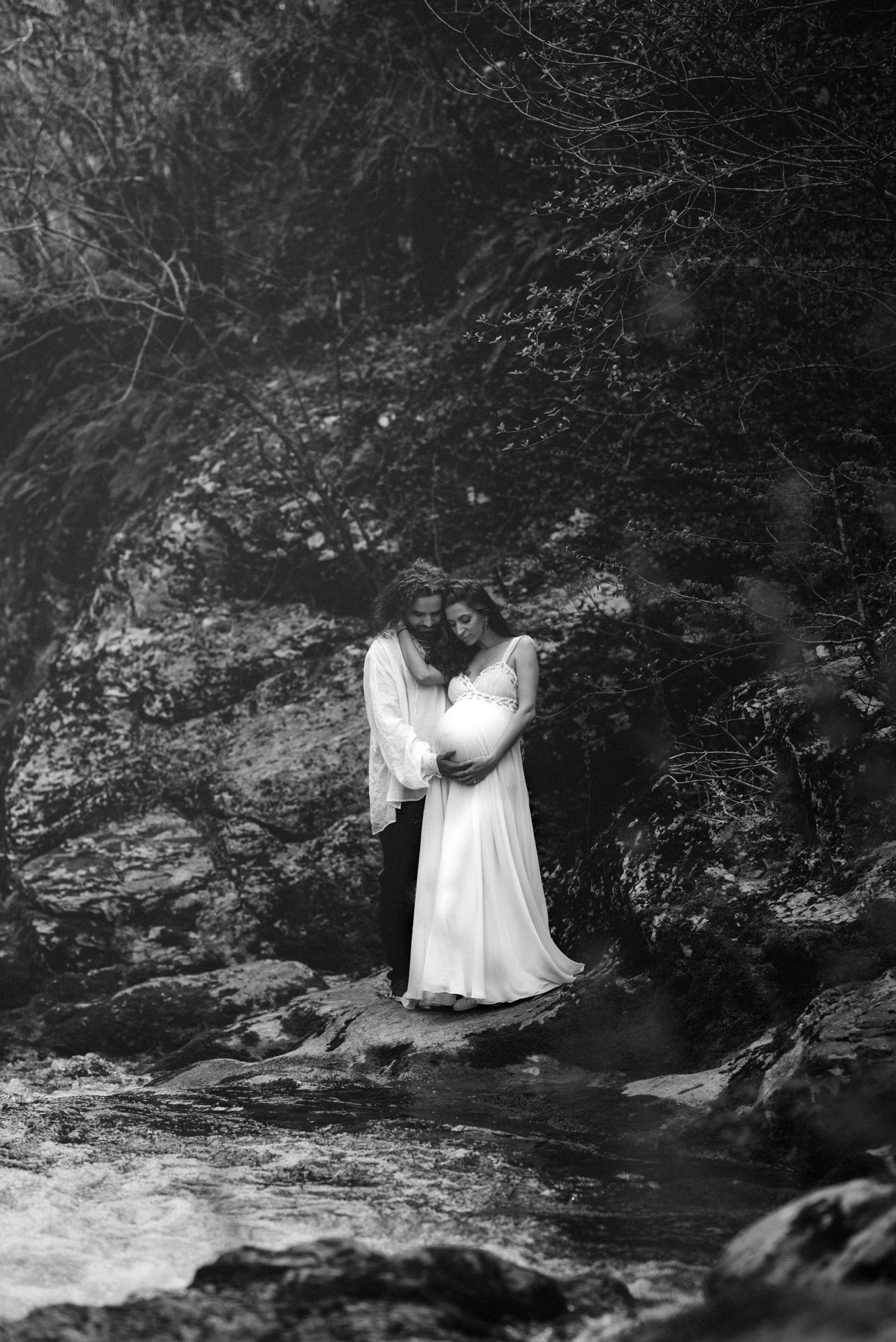 Black and white photo of a couple embracing by a rocky stream, the woman in a white dress cradling her pregnant belly.