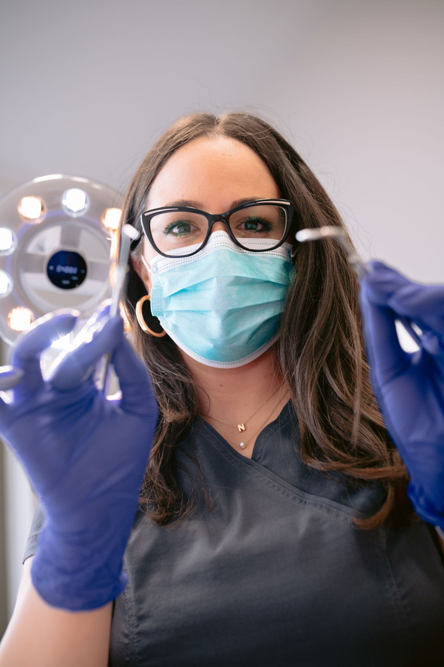 Female dentist in scrubs, mask, and gloves holds dental tools under exam light in clinical setting.