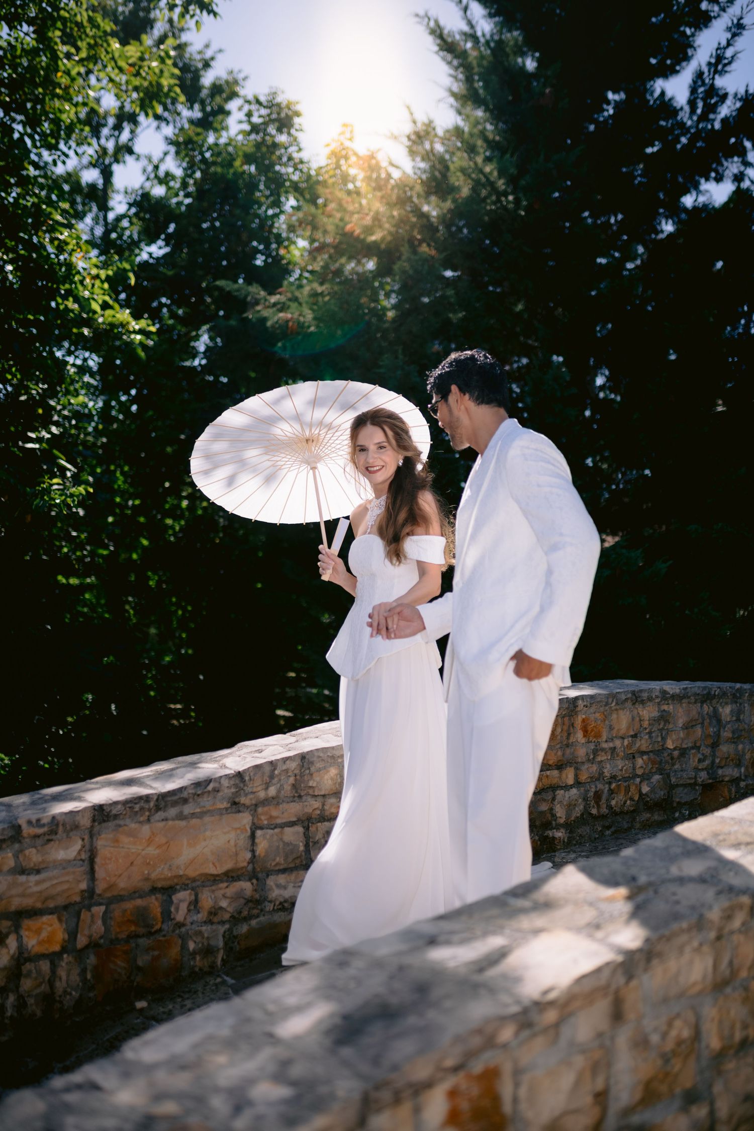 A bride and groom in white attire stand on a stone wall surrounded by lush trees, as she holds a white parasol.