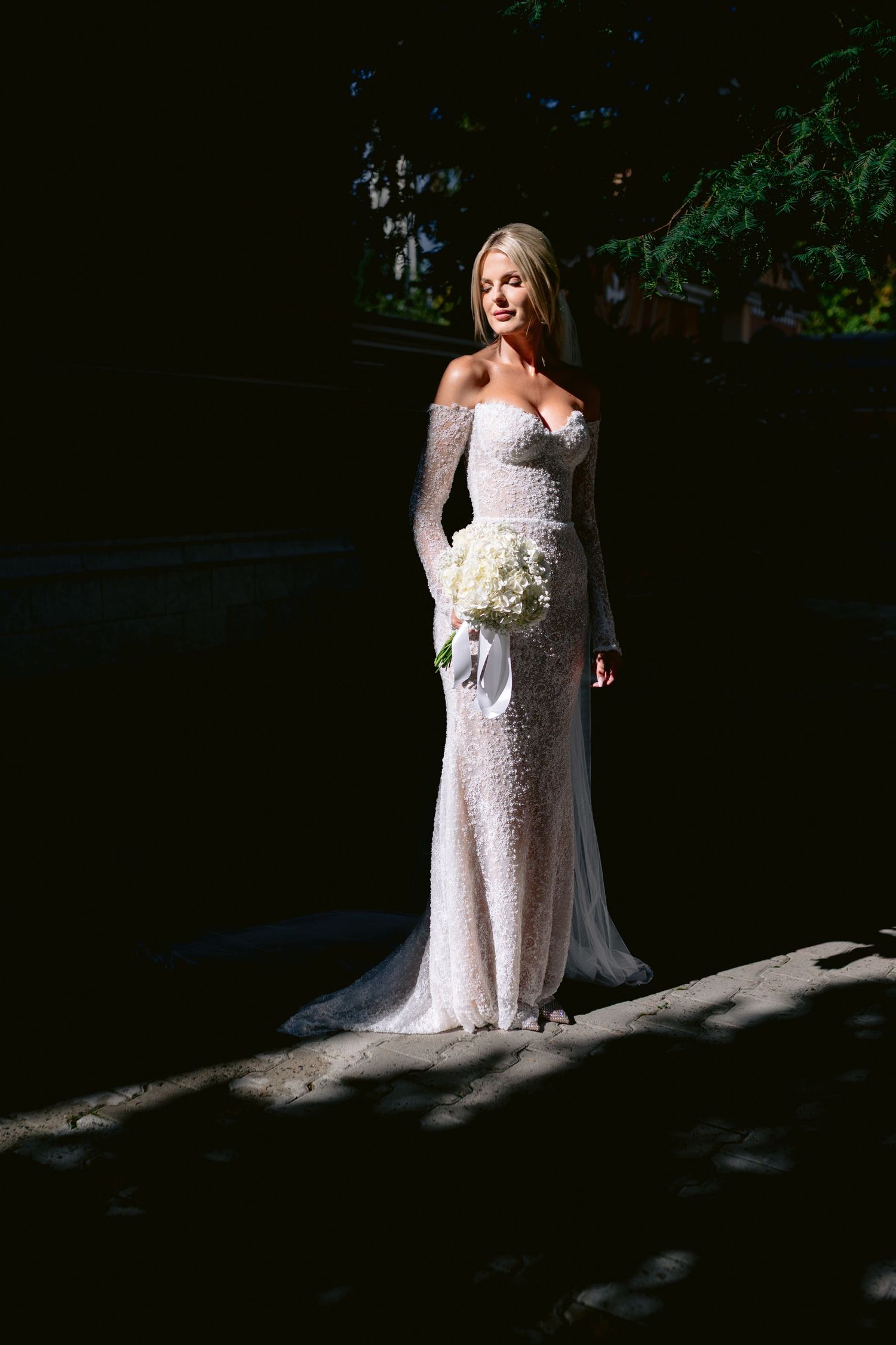 Elegant bride in fitted lace off-shoulder wedding gown holds white bouquet while standing in dramatic lighting.