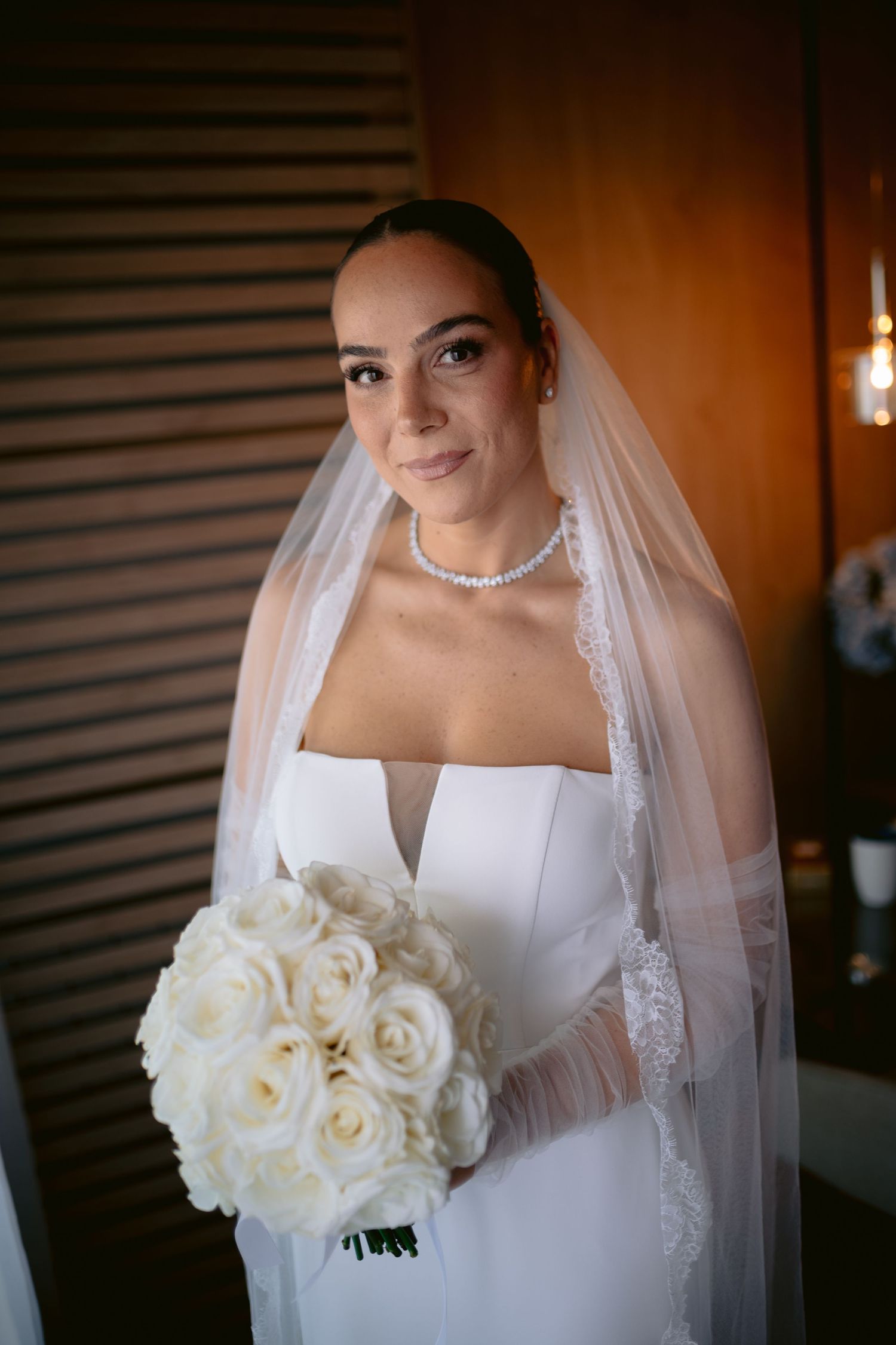 A bride in a white wedding dress holds a bouquet of cream roses against a warm, dimly lit background.