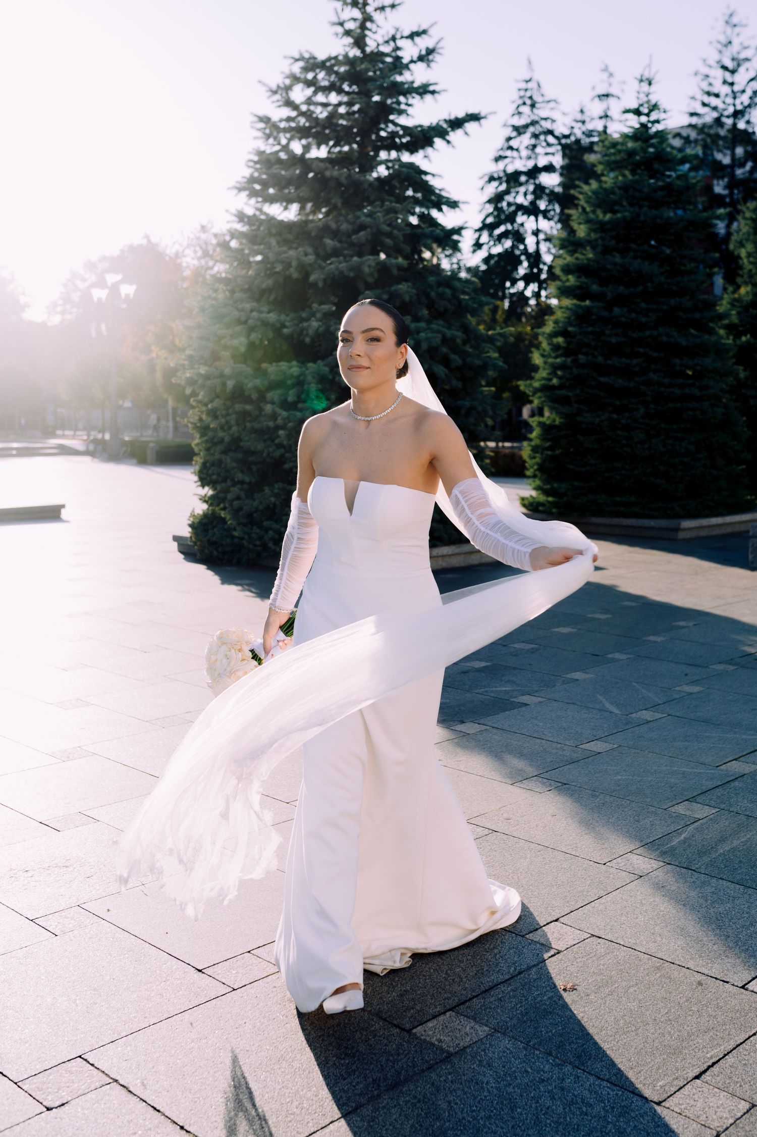A bride in an elegant strapless white wedding gown with long gloves poses gracefully outdoors among evergreen trees.