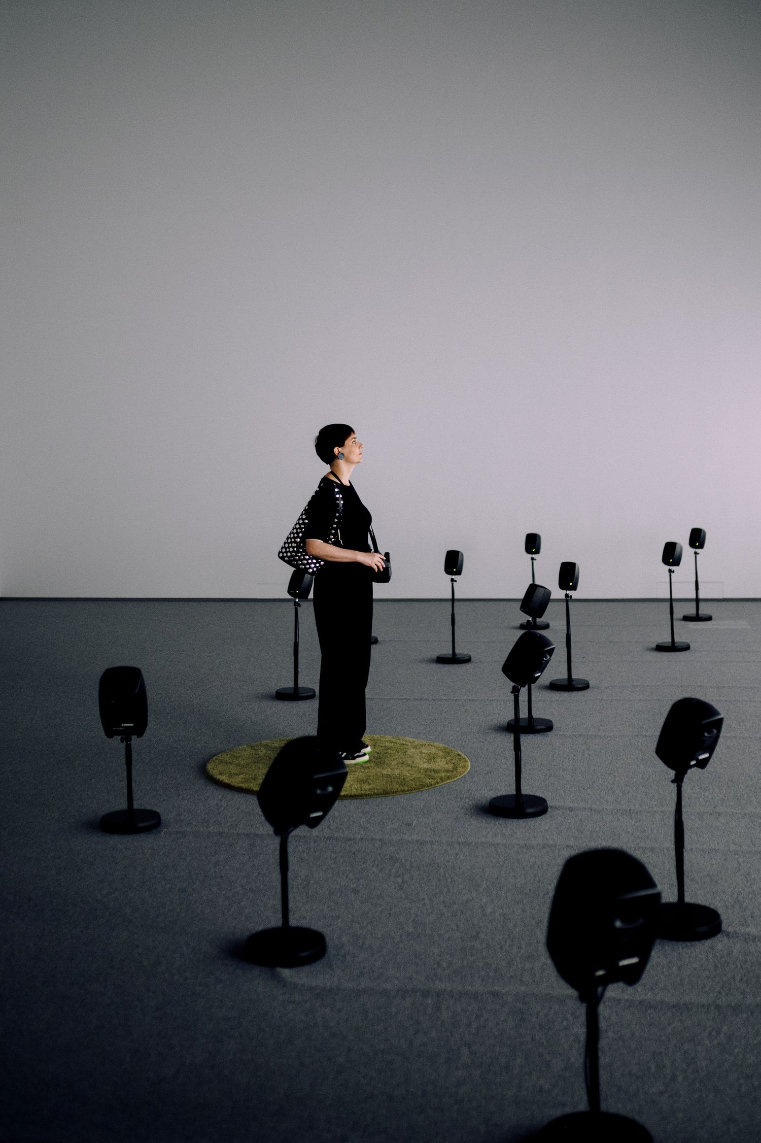 A person stands in a minimalist art installation surrounded by black speaker stands on a gray floor.