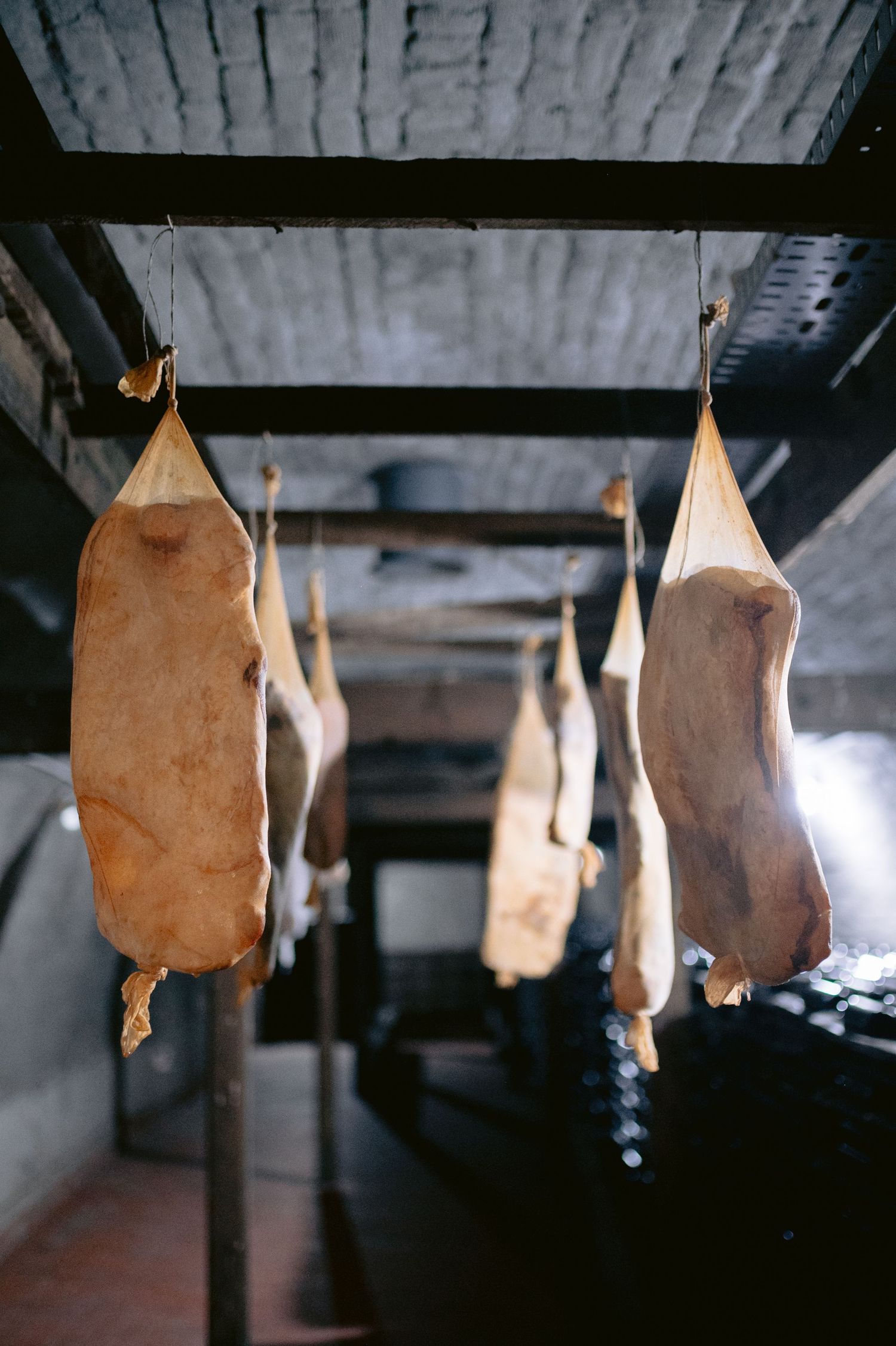 Several cured meat or ham legs hanging from hooks in a dark cellar storage room.
