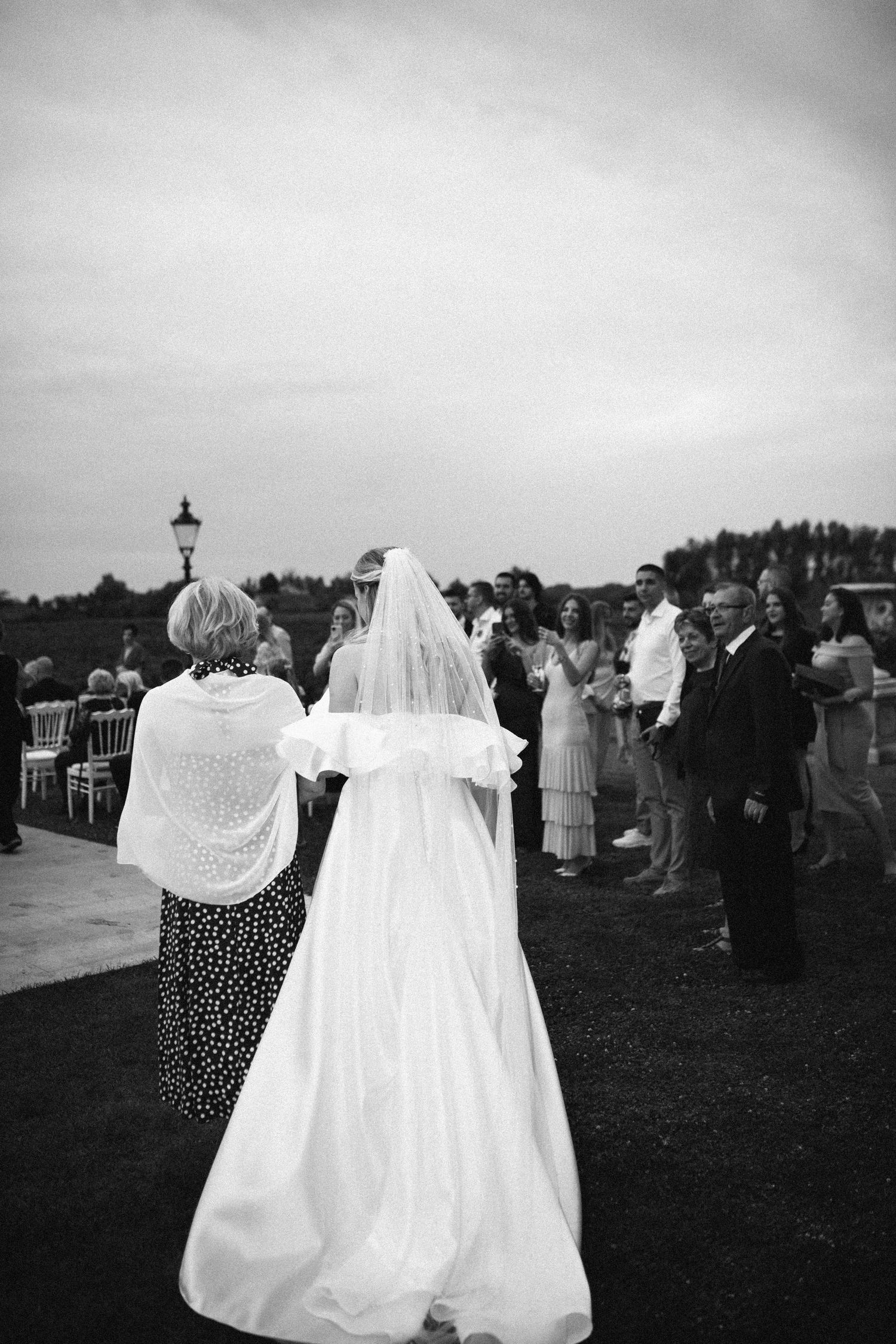 A black and white panoramic sequence of wedding guests gathered outdoors with a bride in a flowing white gown.