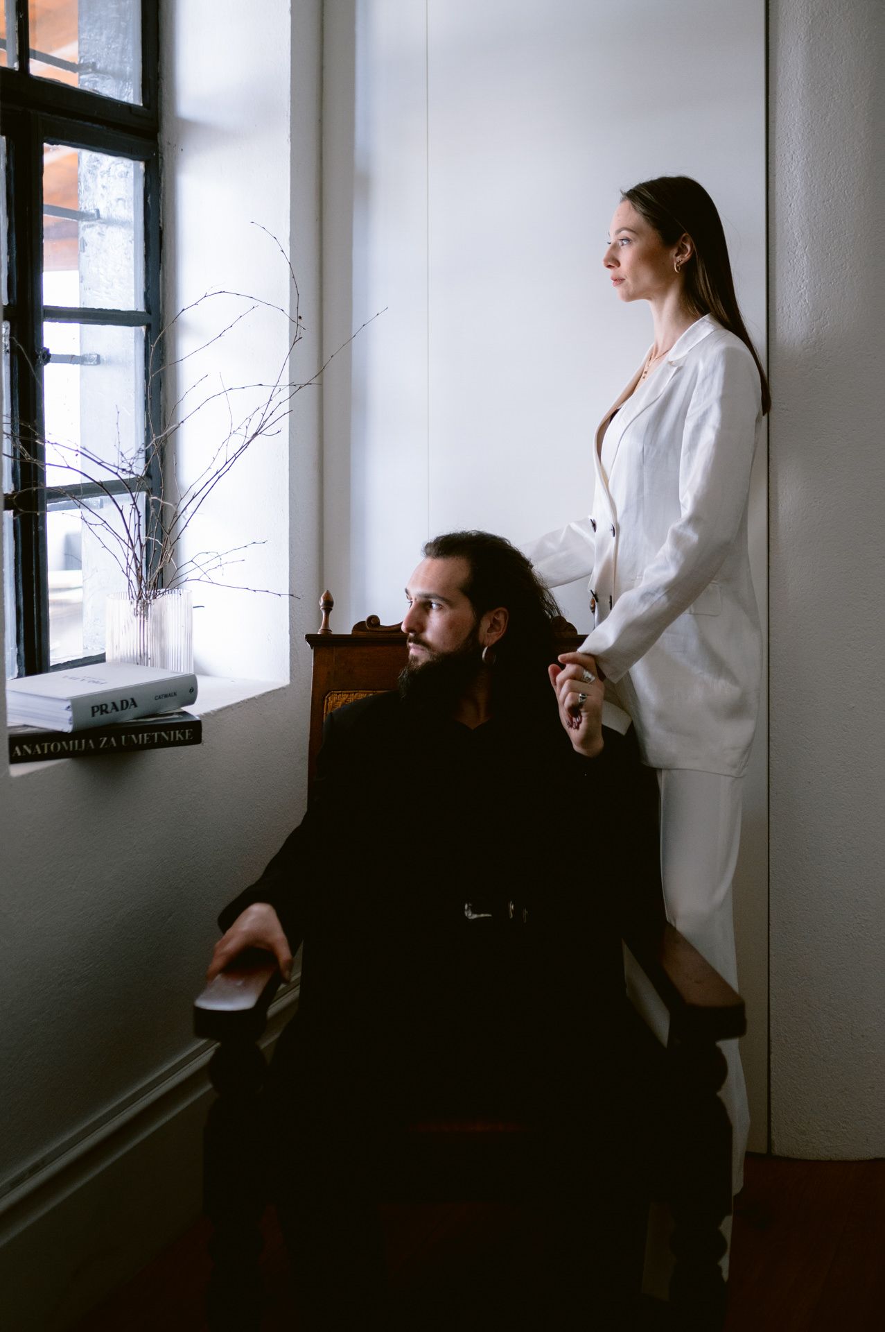 Two people in contrasting black and white outfits pose artistically by a window in moody lighting.