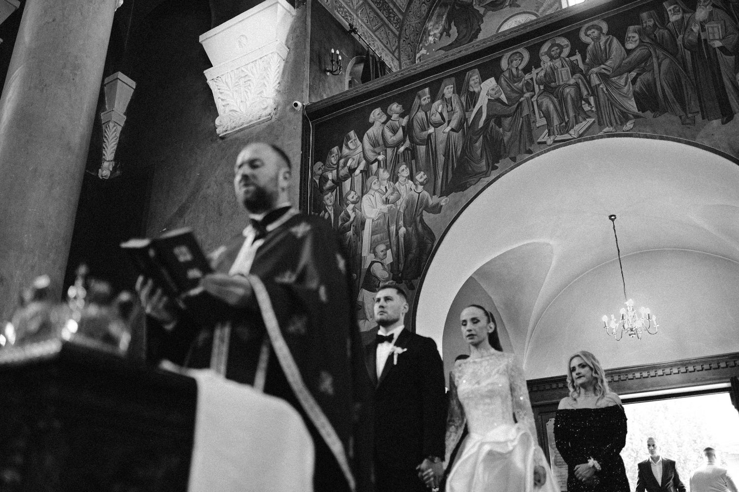 Black and white photo of a couple exchanging vows in an ornate Orthodox church with a priest reading from a book.