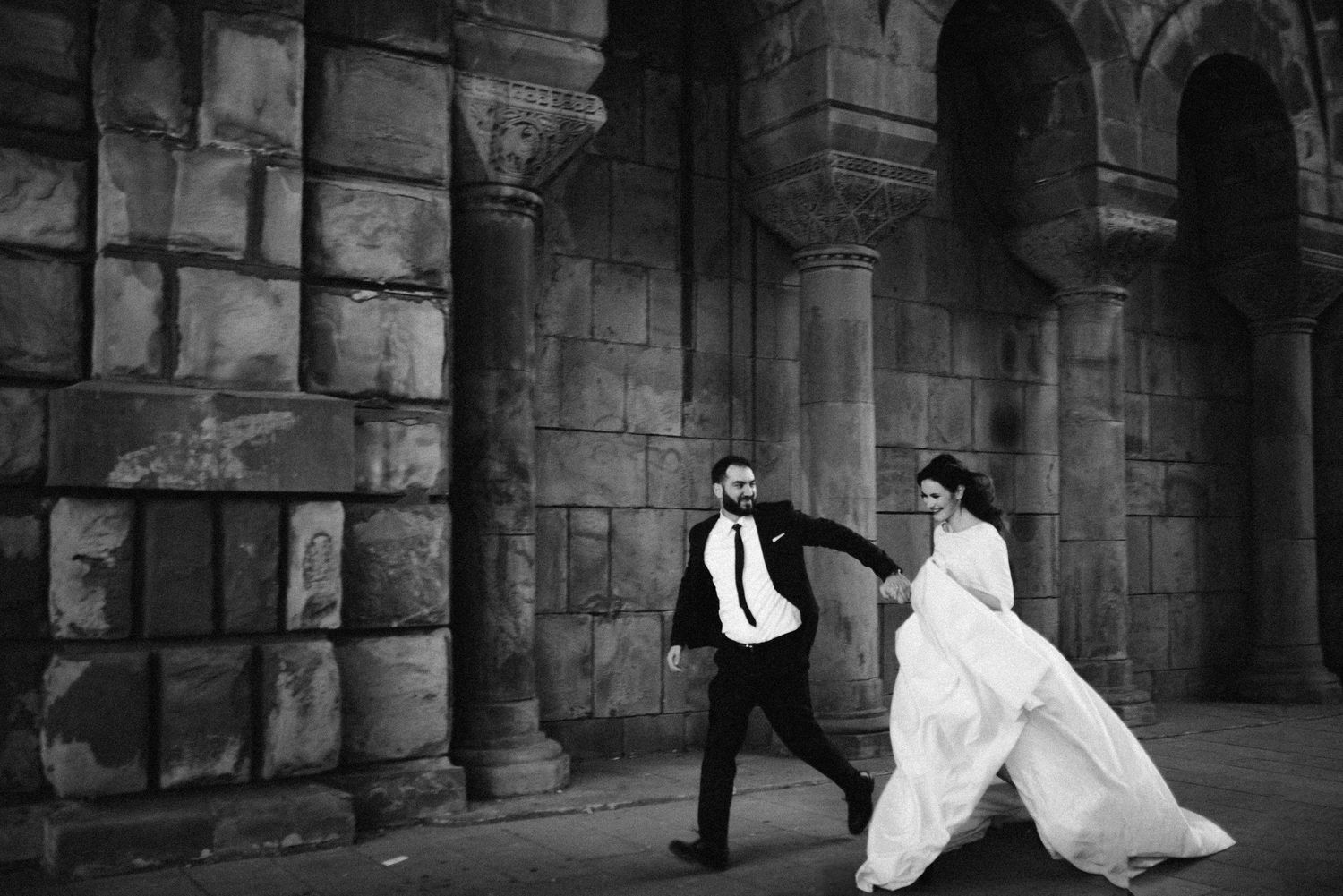 Black and white photo of bride and groom walking hand in hand outside a historic stone building.