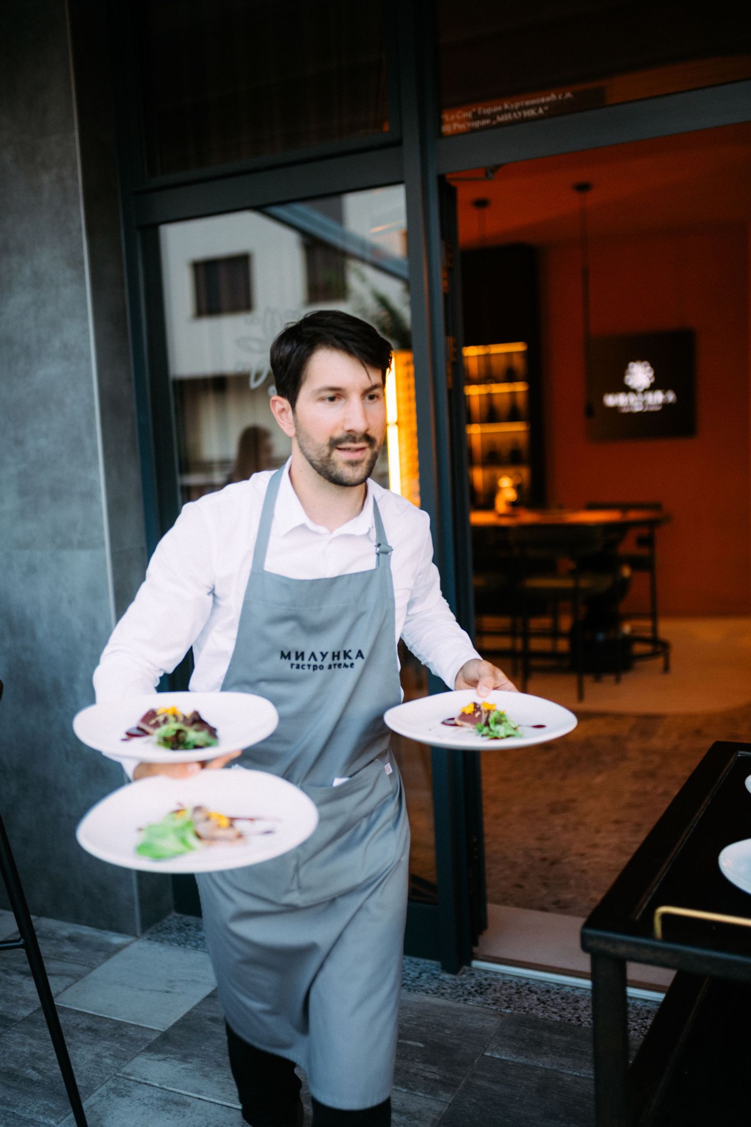 Smiling waiter in gray apron carries three elegant plated dishes outside a modern restaurant entrance.