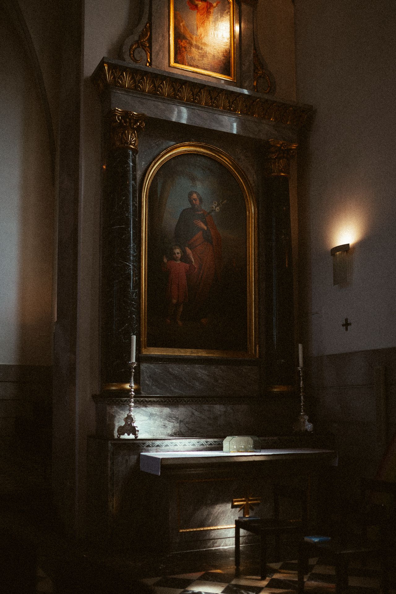 Dark church altar with ornate golden frame painting of saint and child, candlestick, and warm wall sconce lighting.