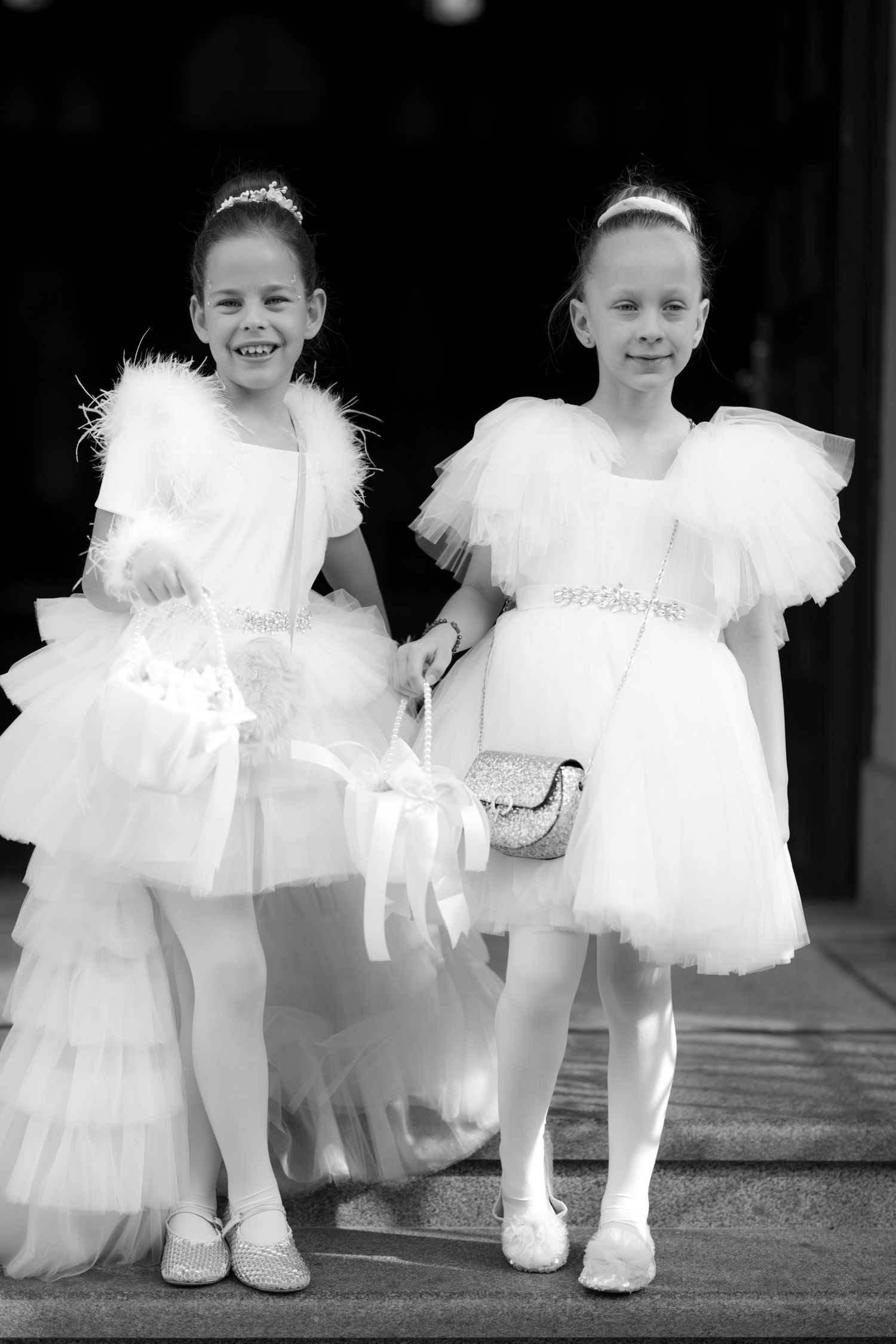 Two young flower girls in white tulle dresses holding baskets pose together in a black and white photo.