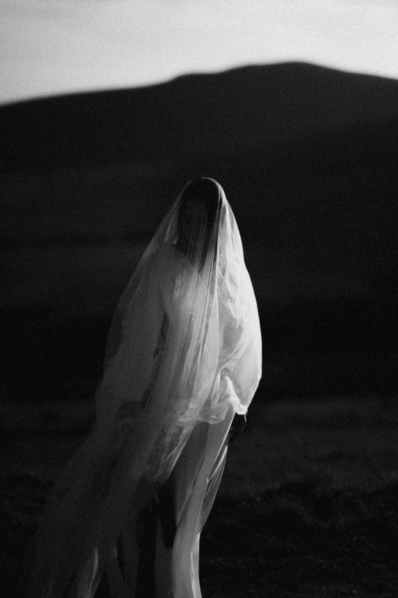 Black and white photo of a veiled bride standing in a dramatic dark landscape with rolling hills.