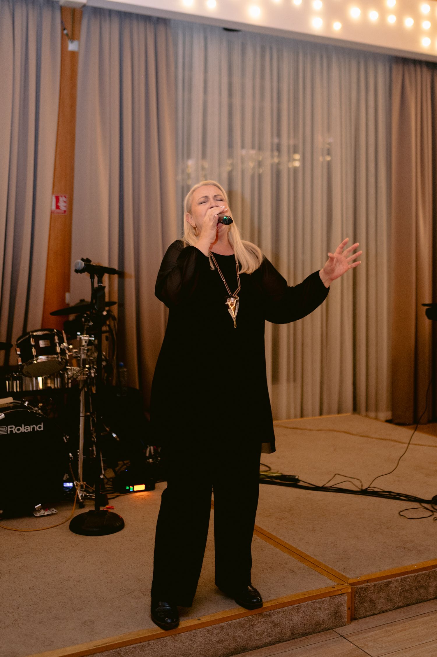 Blonde female singer performing passionately in black outfit on stage with Roland drum kit at wedding reception.
