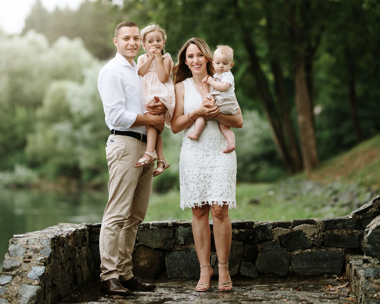 A smiling family of four poses outdoors near a stone wall and pond, parents holding two young children in white outfits.