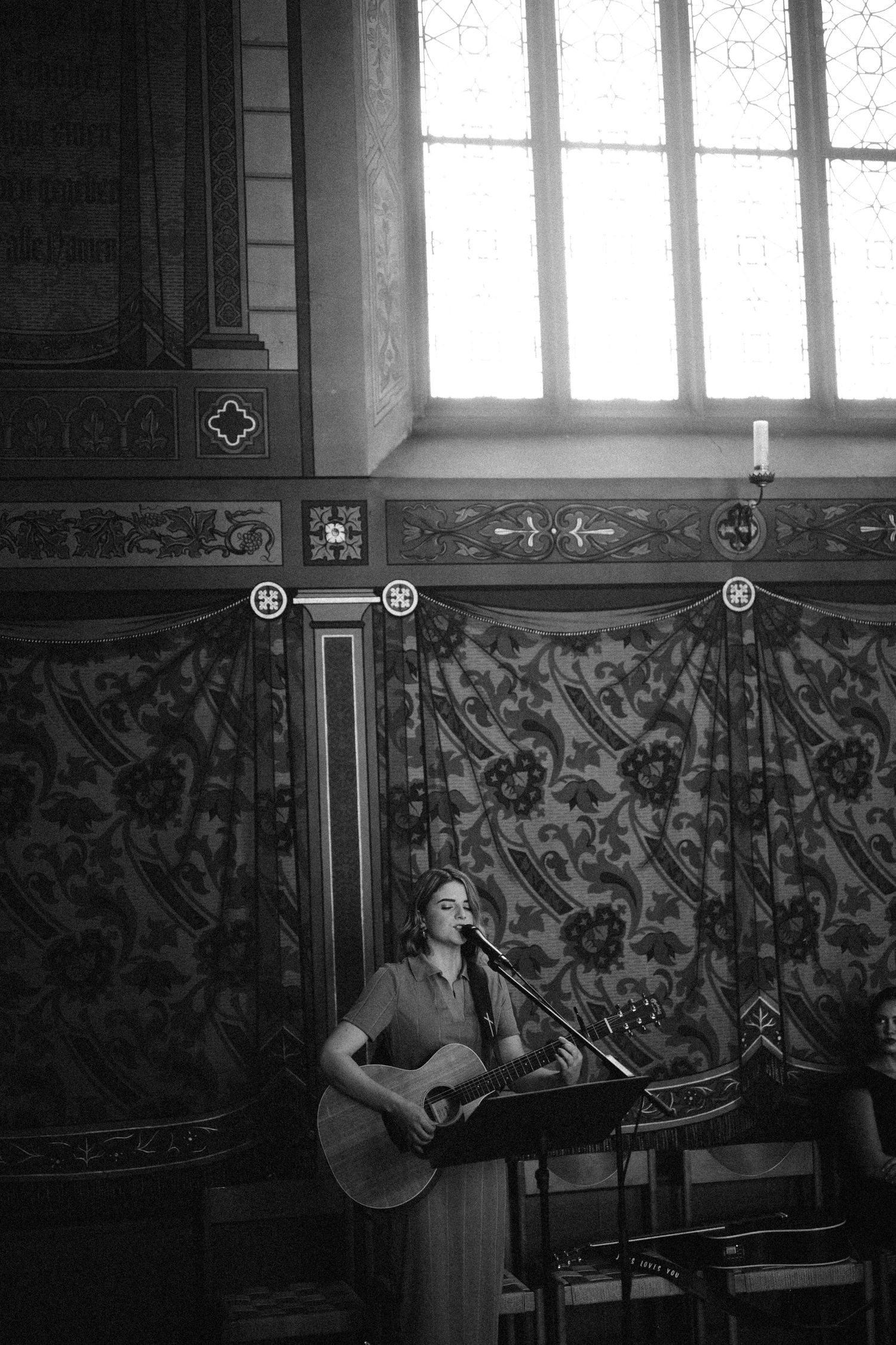 Woman playing acoustic guitar and singing into a microphone in an ornate church setting, black and white photo.