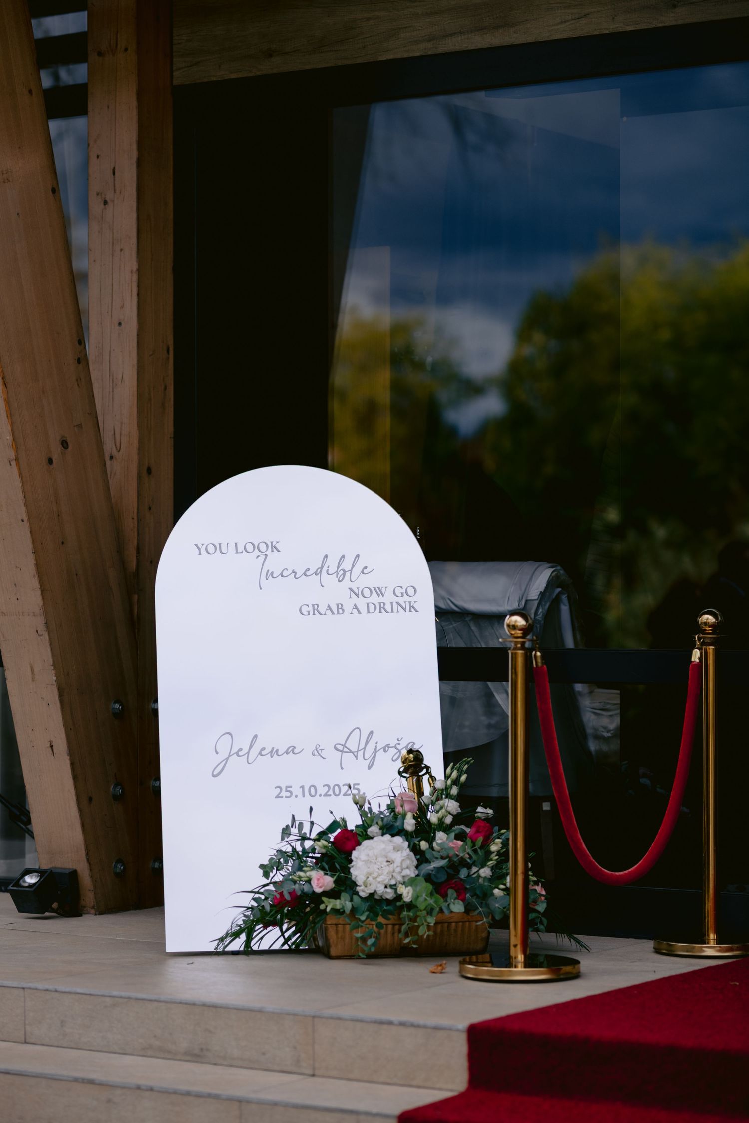 Elegant white arched wedding welcome sign with floral arrangement and red velvet rope barriers at venue entrance.