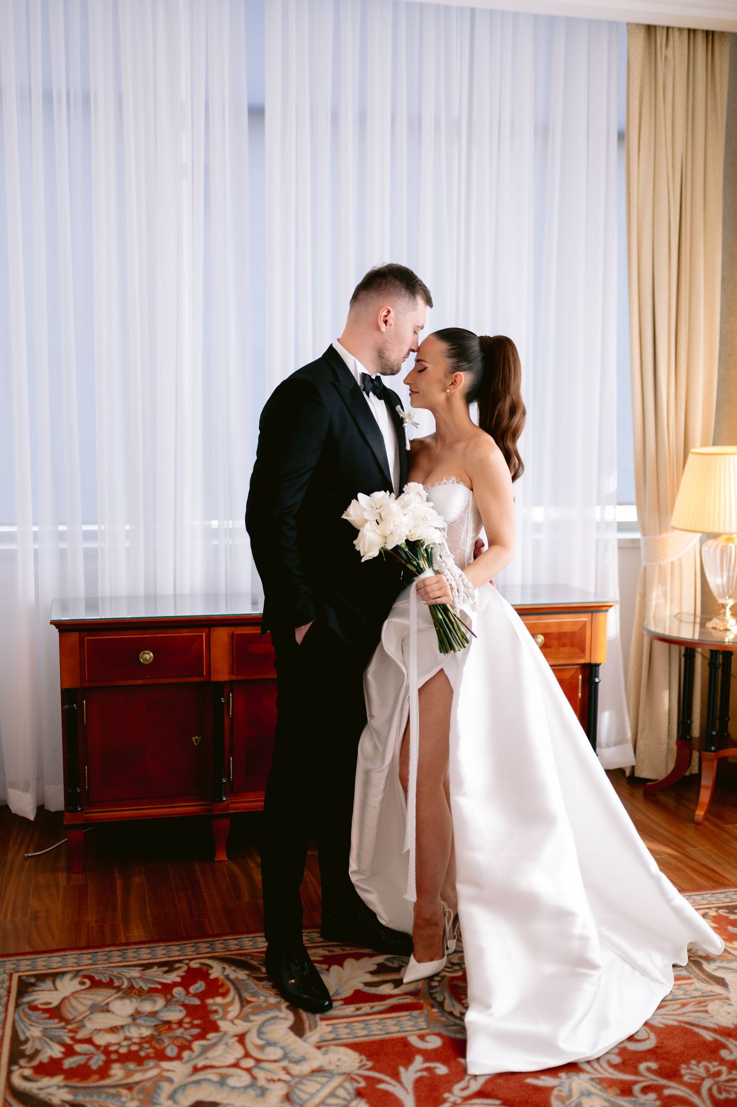 Romantic wedding couple sharing a dance dip in an elegant hotel room with red carpeting and sheer curtains.