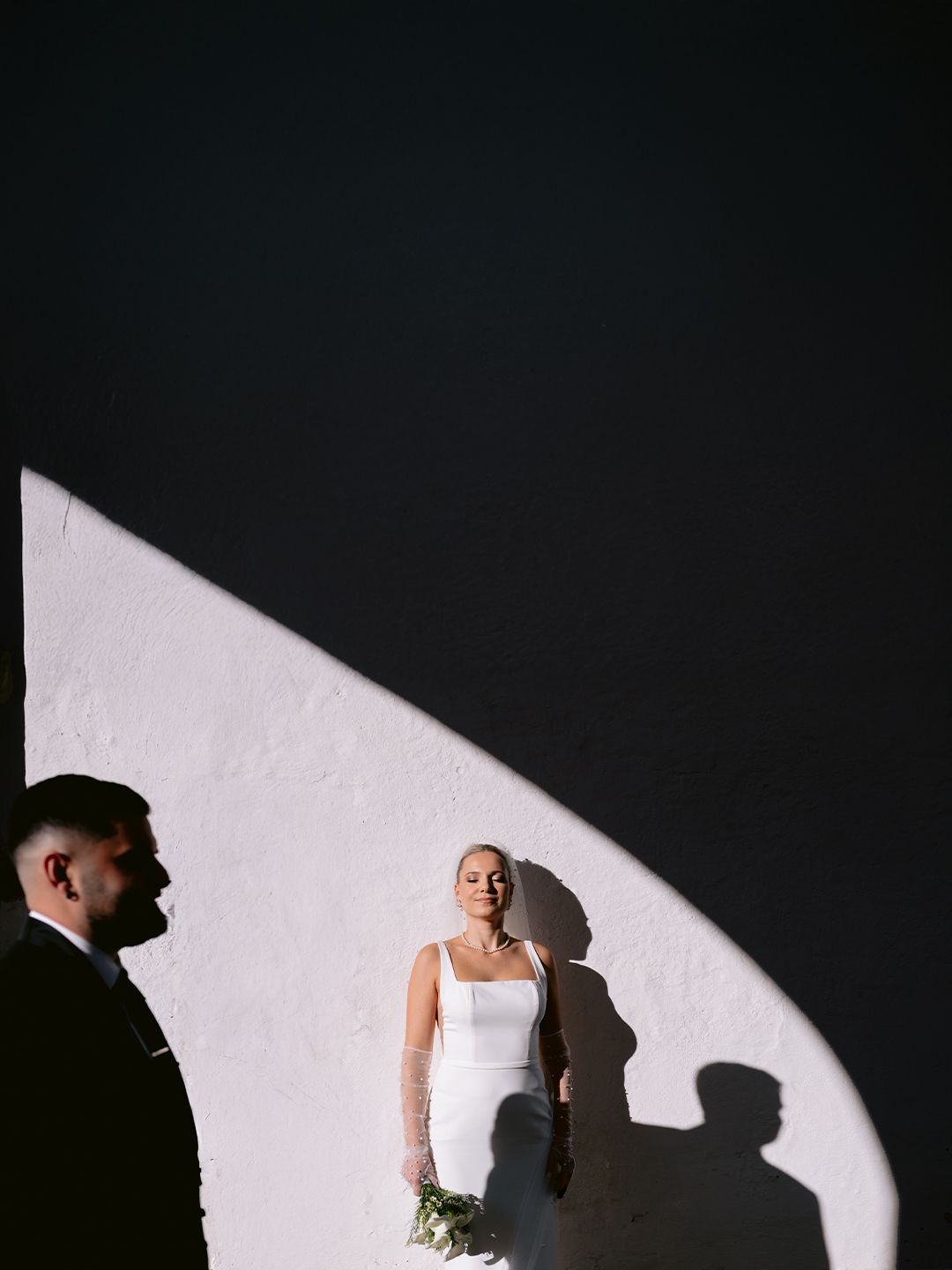 Dramatic wedding photo with stark lighting showing silhouettes against a white wall, with bouquet and elegant white dress.