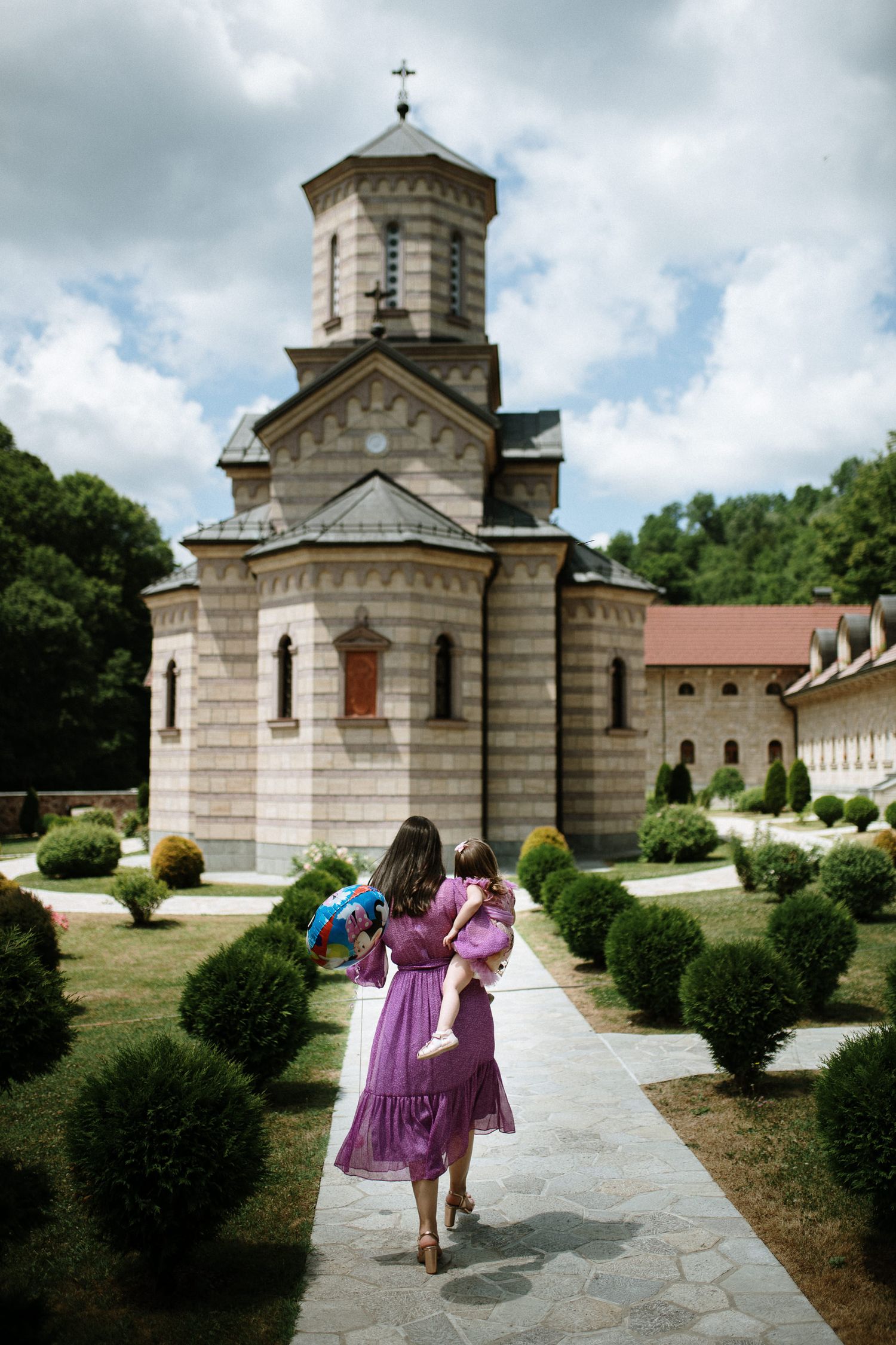 Woman in purple dress holding child walks toward ornate Orthodox monastery church surrounded by manicured gardens.