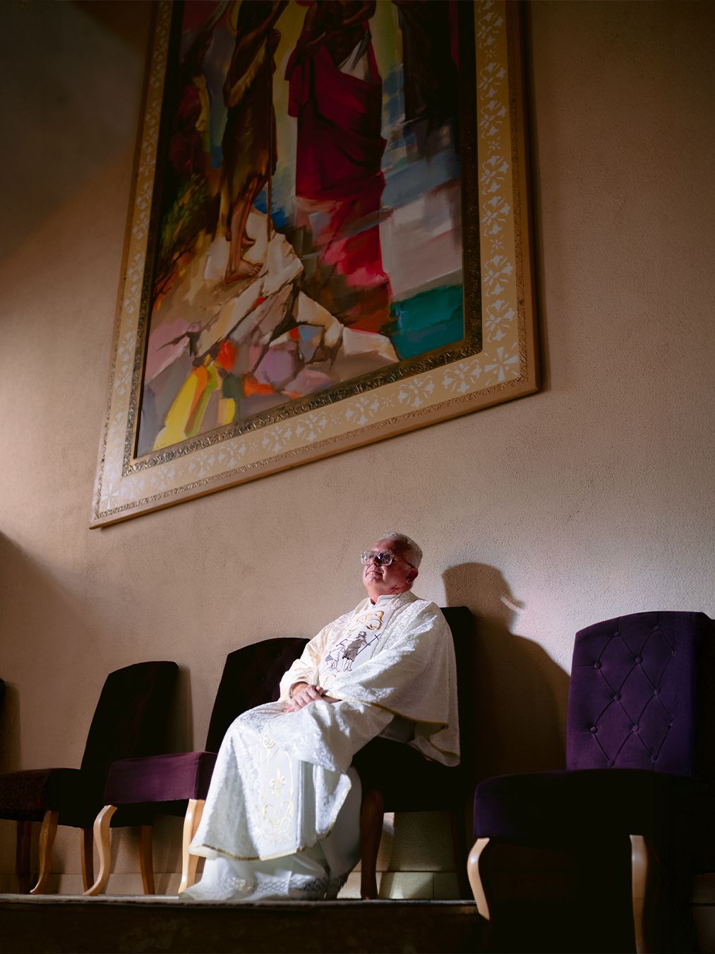 A robed clergy member sits beneath a colorful abstract painting in a dimly lit room with purple chairs.