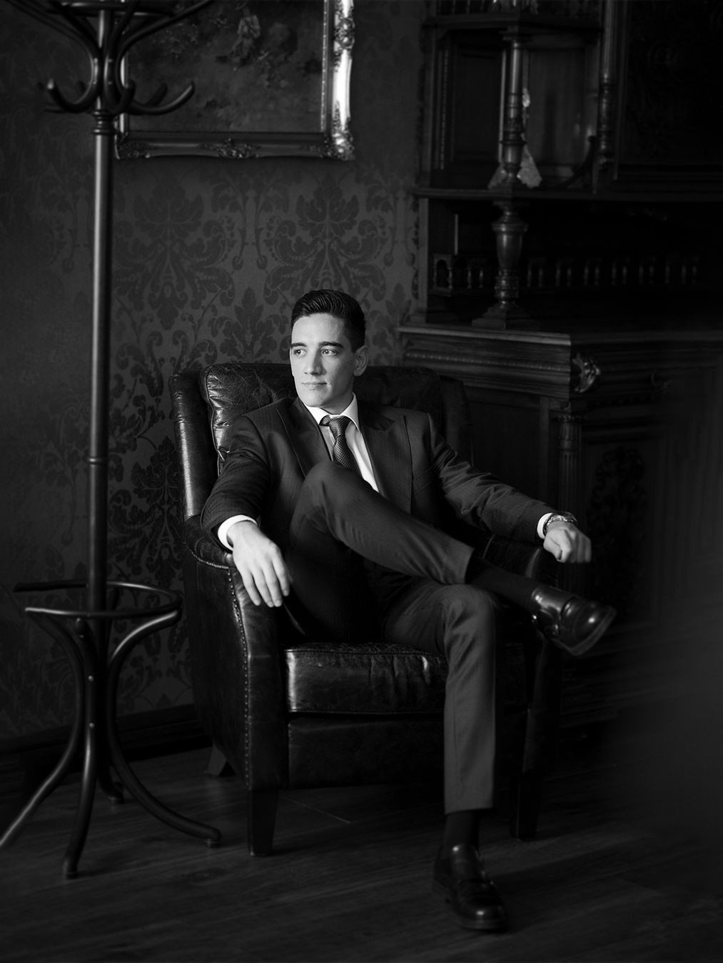 Man in suit sitting confidently in leather chair, black and white portrait photography in elegant room.