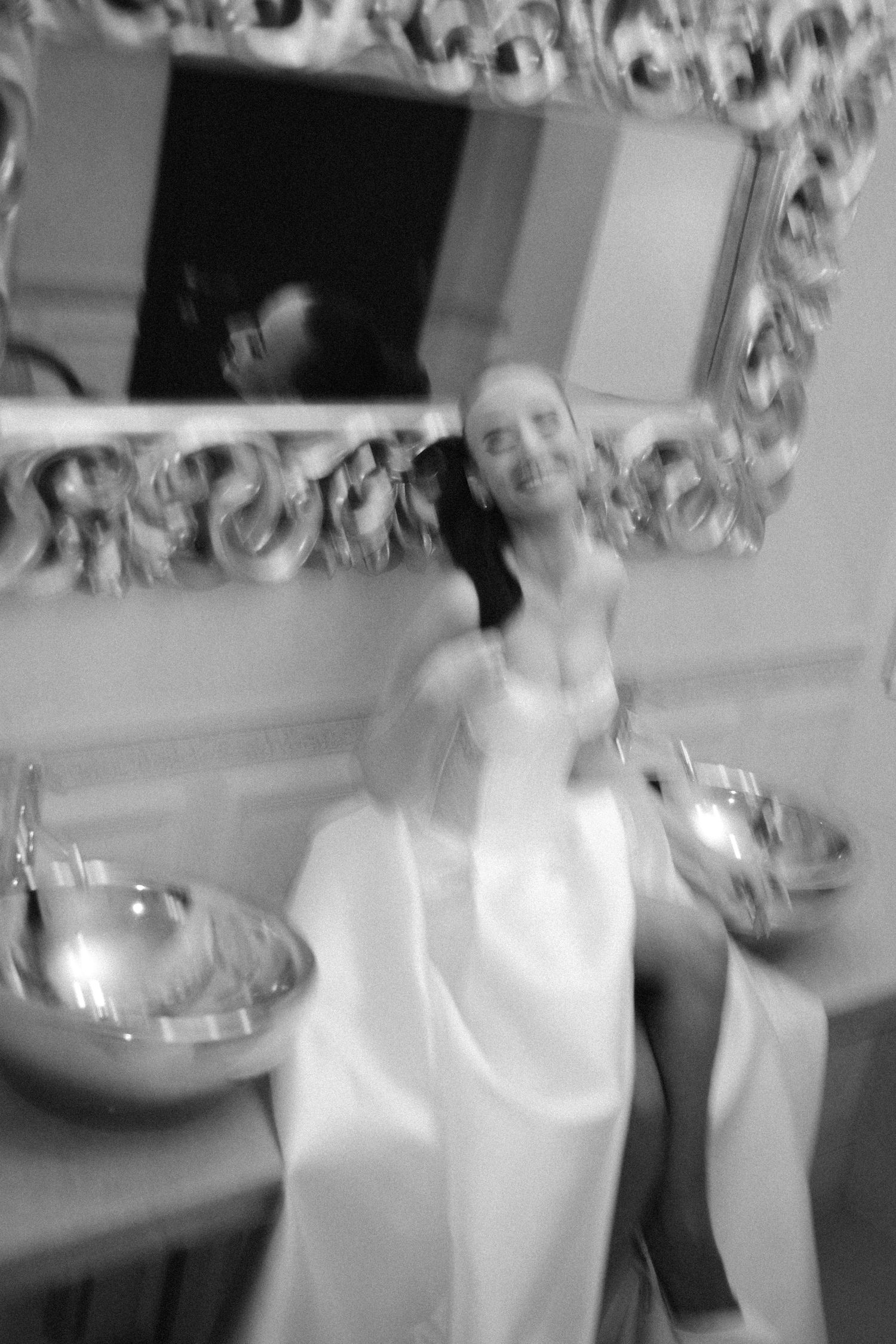Blurred black and white photo of a bride in white dress sitting between two ornate sinks below a decorative mirror.