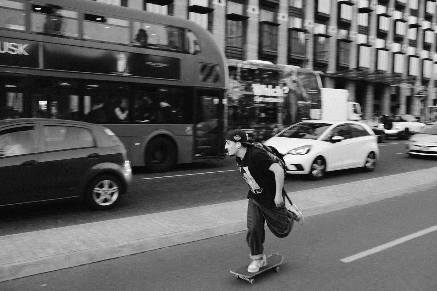 A person rides a skateboard down a city street lined with parked cars and buildings.